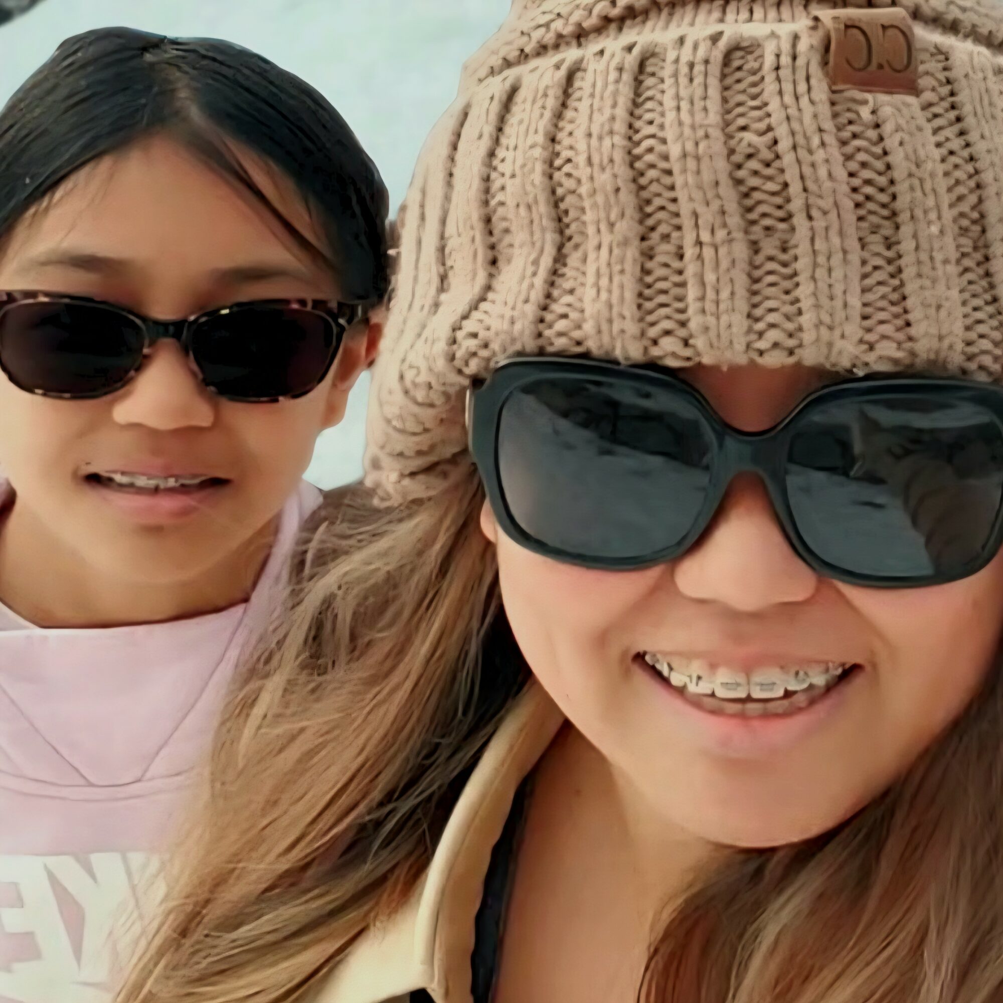 Two smiling women wearing sunglasses, one with a beige knit hat, outdoors.