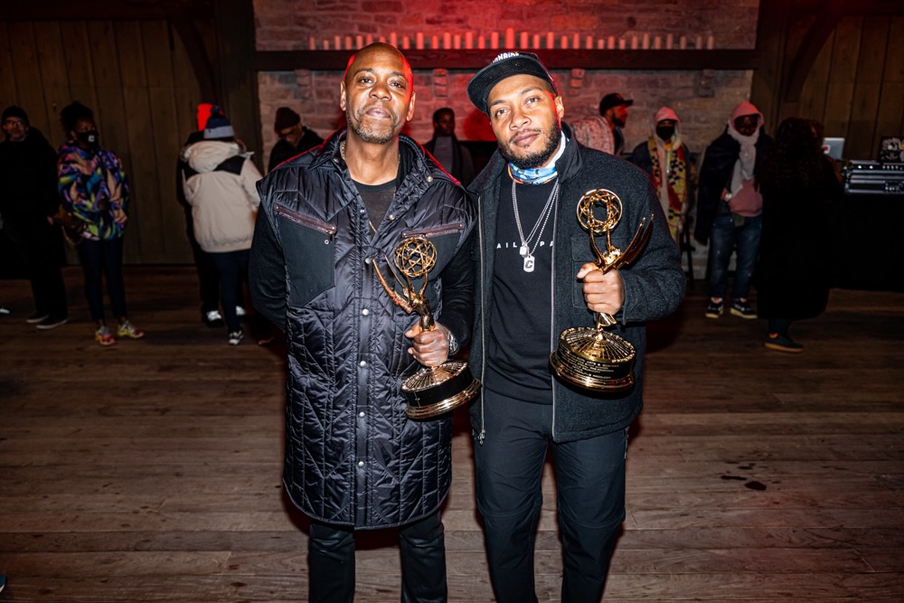 Two men holding trophies, standing indoors with people in the background, smiling at the camera.