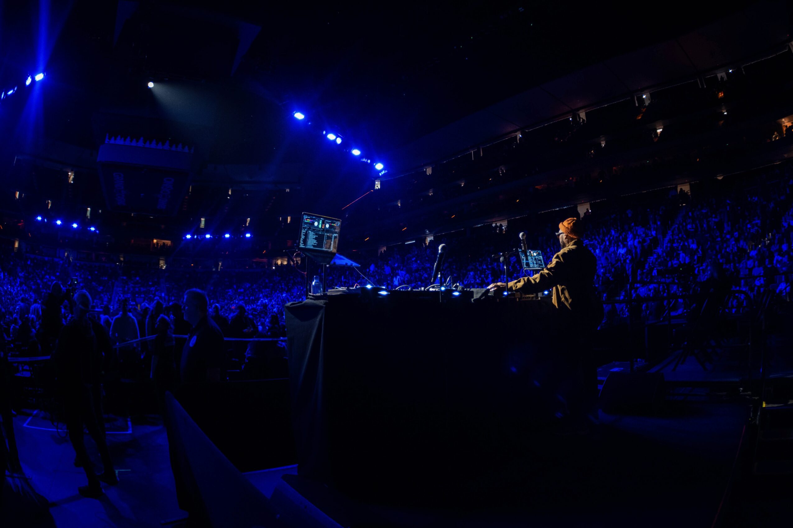 DJ performing on stage with blue lighting, audience in a large indoor venue, screens displaying visuals, dark atmosphere.