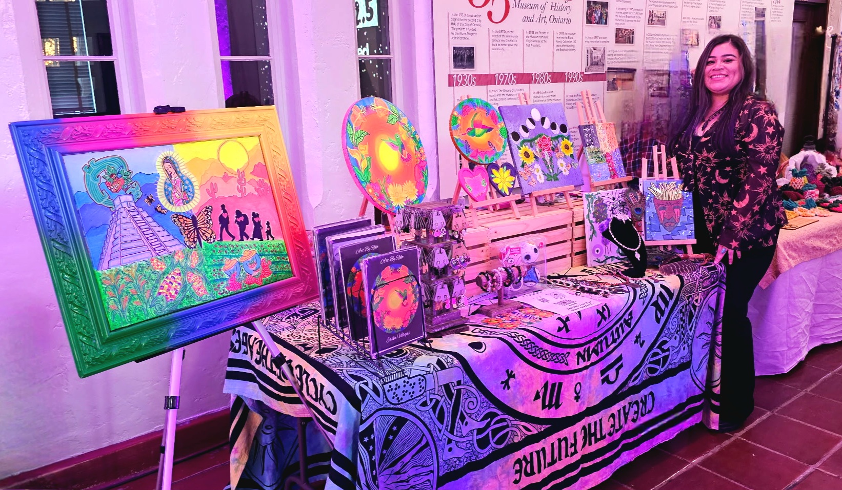 Woman standing next to a table with colorful artwork and decorative items, inside a room with newspaper-covered walls.