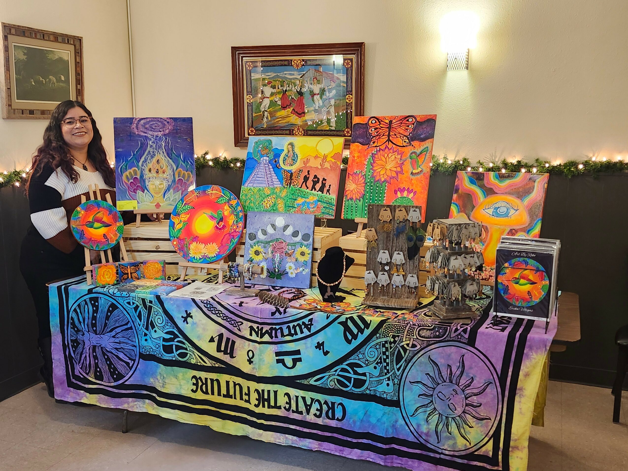 Woman standing next to colorful paintings and crafts on a table at an indoor event.