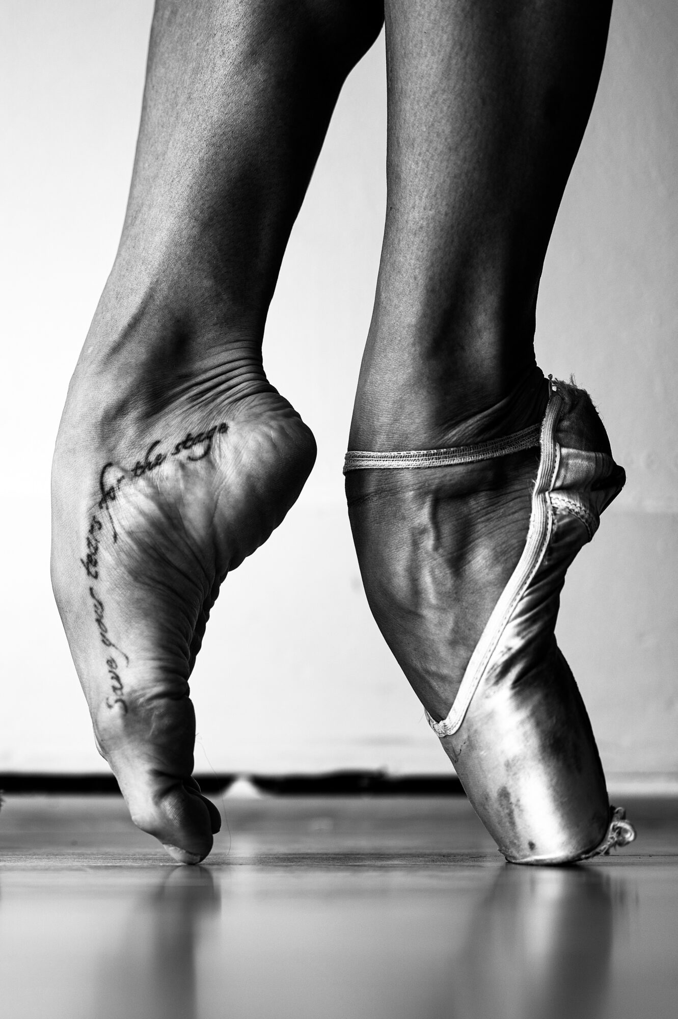 Close-up of ballet dancer's feet in pointe shoes, one foot on tiptoe, the other flat, with tattoo on ankle.