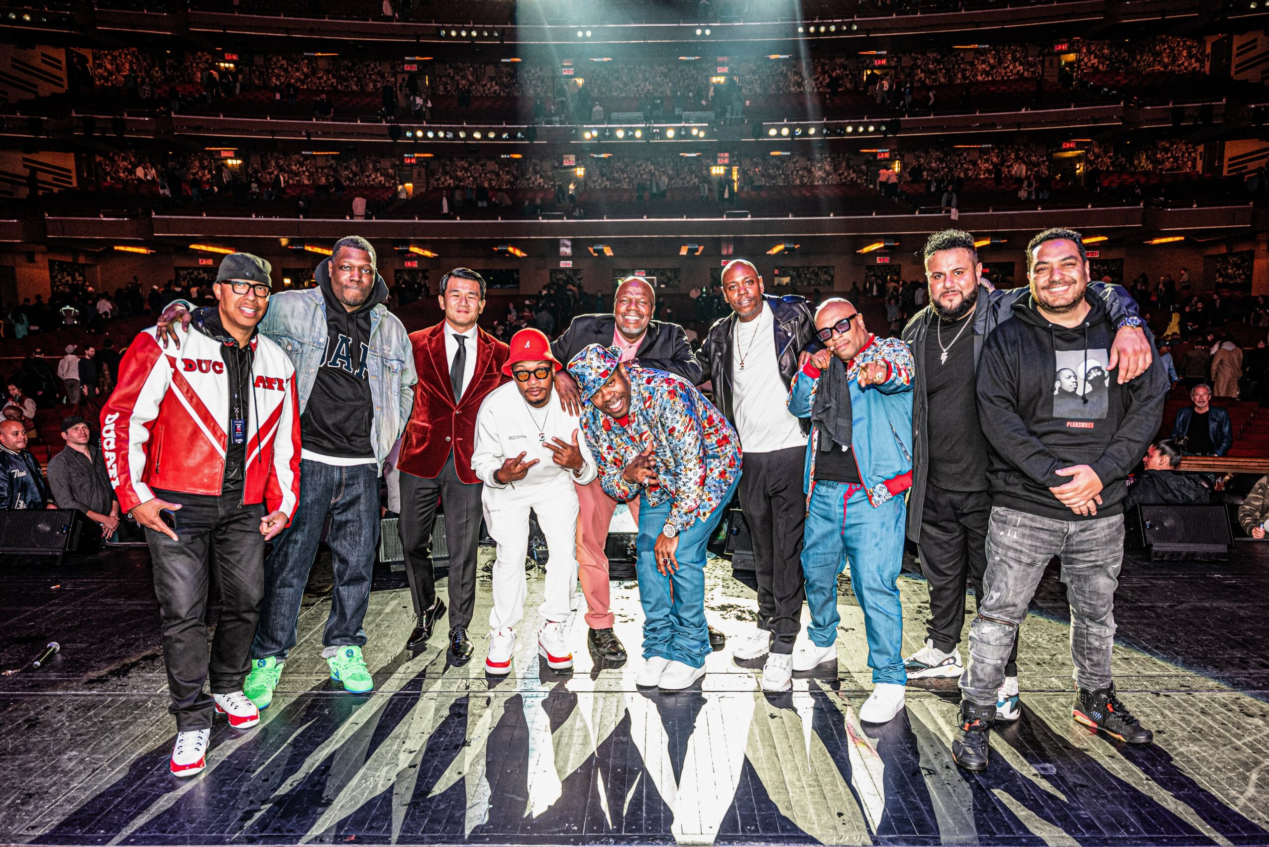 Group of ten people standing on stage in a large indoor arena with audience in background.