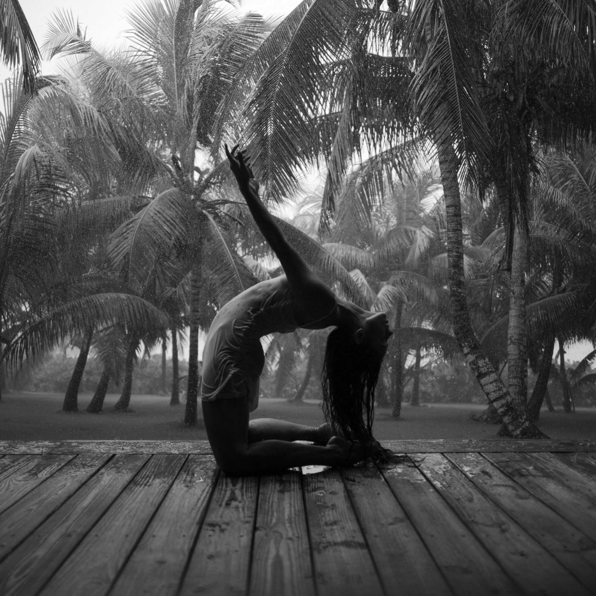 Person practicing yoga on wooden deck with palm trees in background, performing a backbend pose with one arm raised.