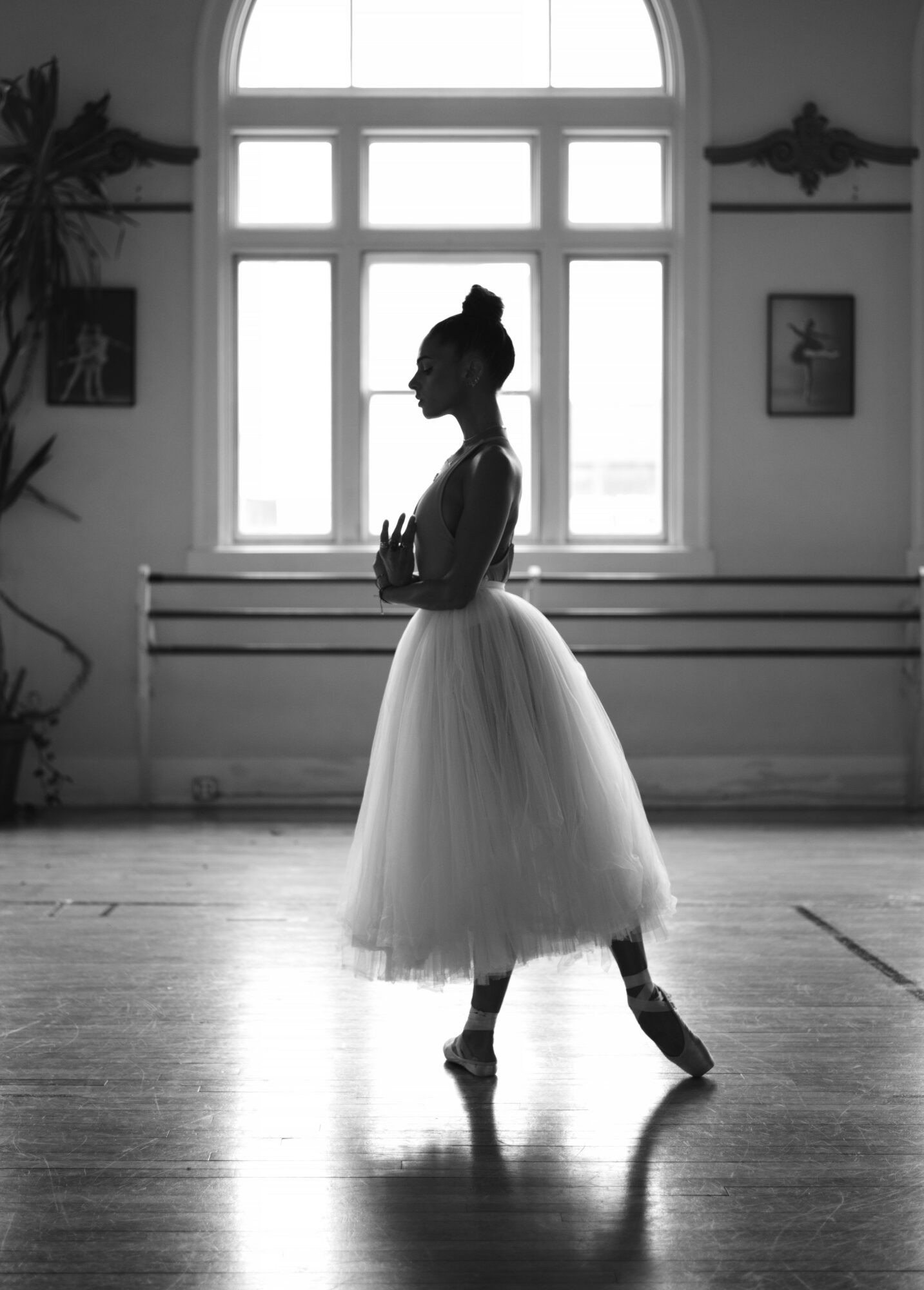 Silhouette of a ballerina in a tutu and ballet shoes standing on a wooden floor near a large window.