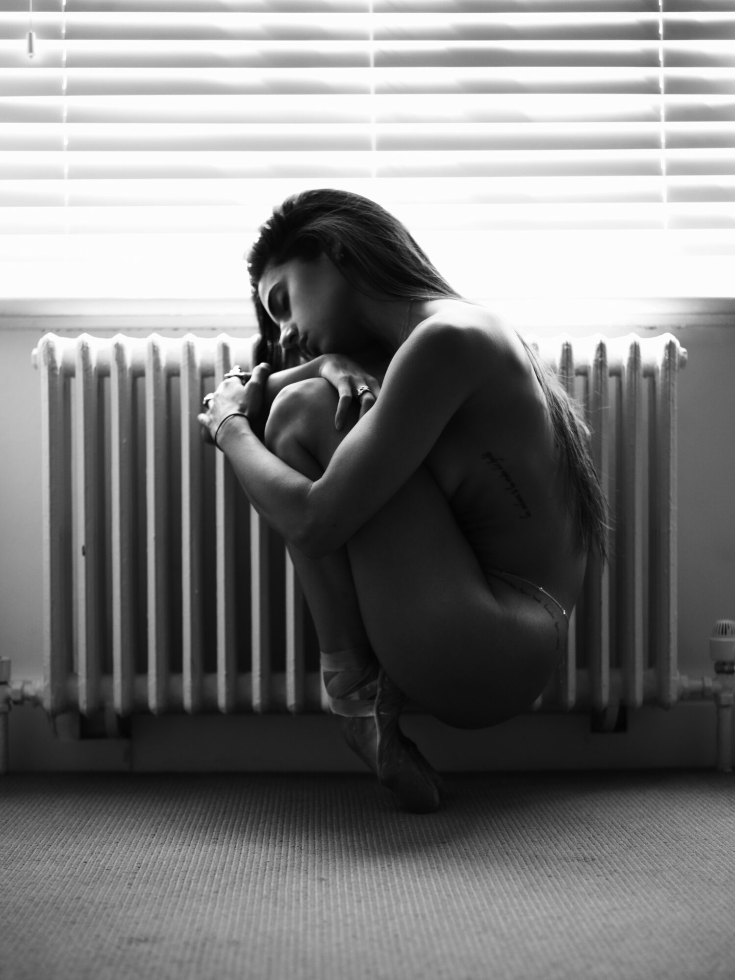Woman sitting on the floor, hugging her knees near a radiator with blinds behind her.