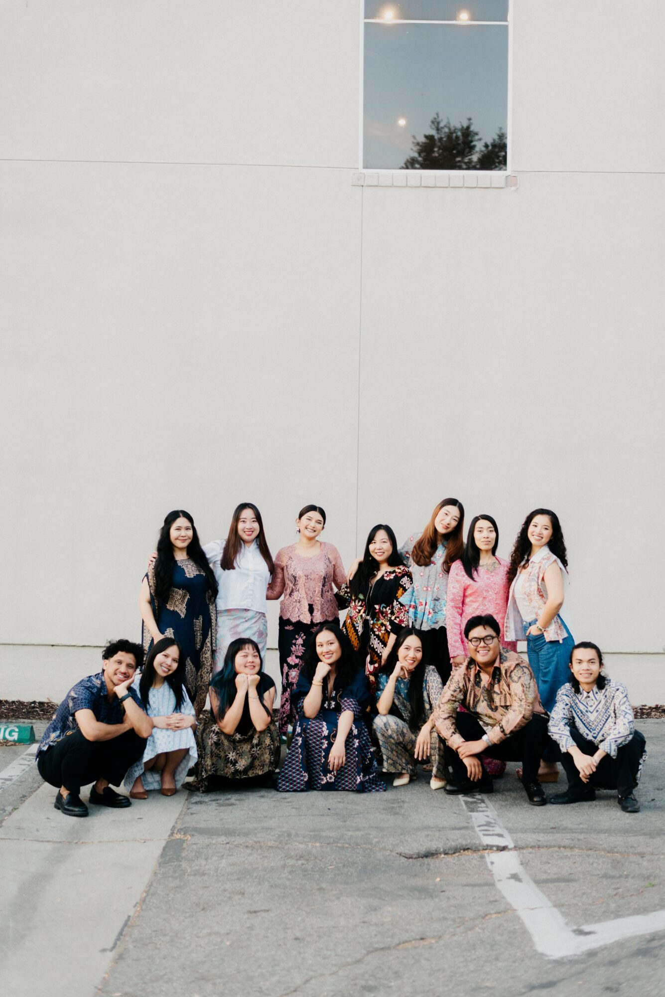 Group of twelve people posing outdoors in front of a white wall with a window, some standing and some kneeling.