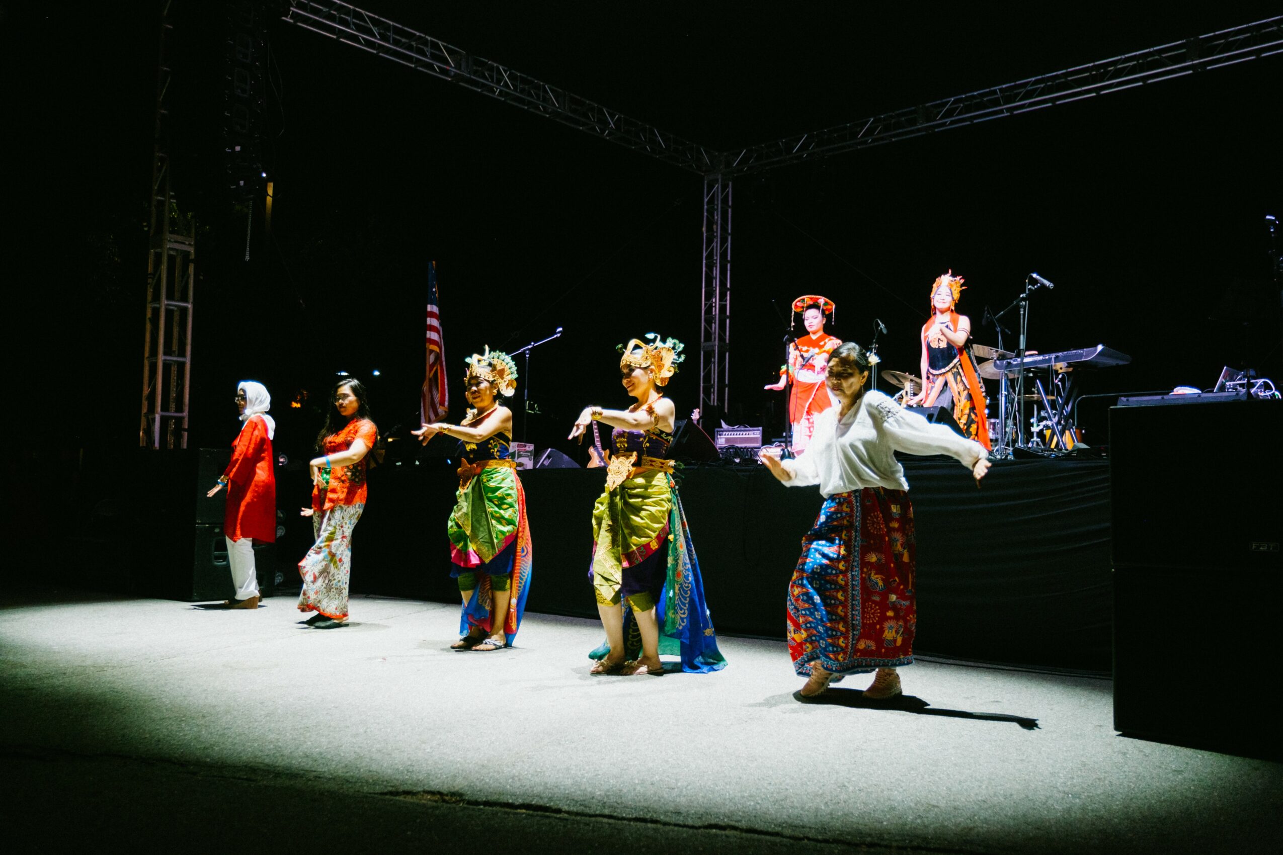 Six performers in traditional costumes dance on stage with musical instruments and equipment in the background.