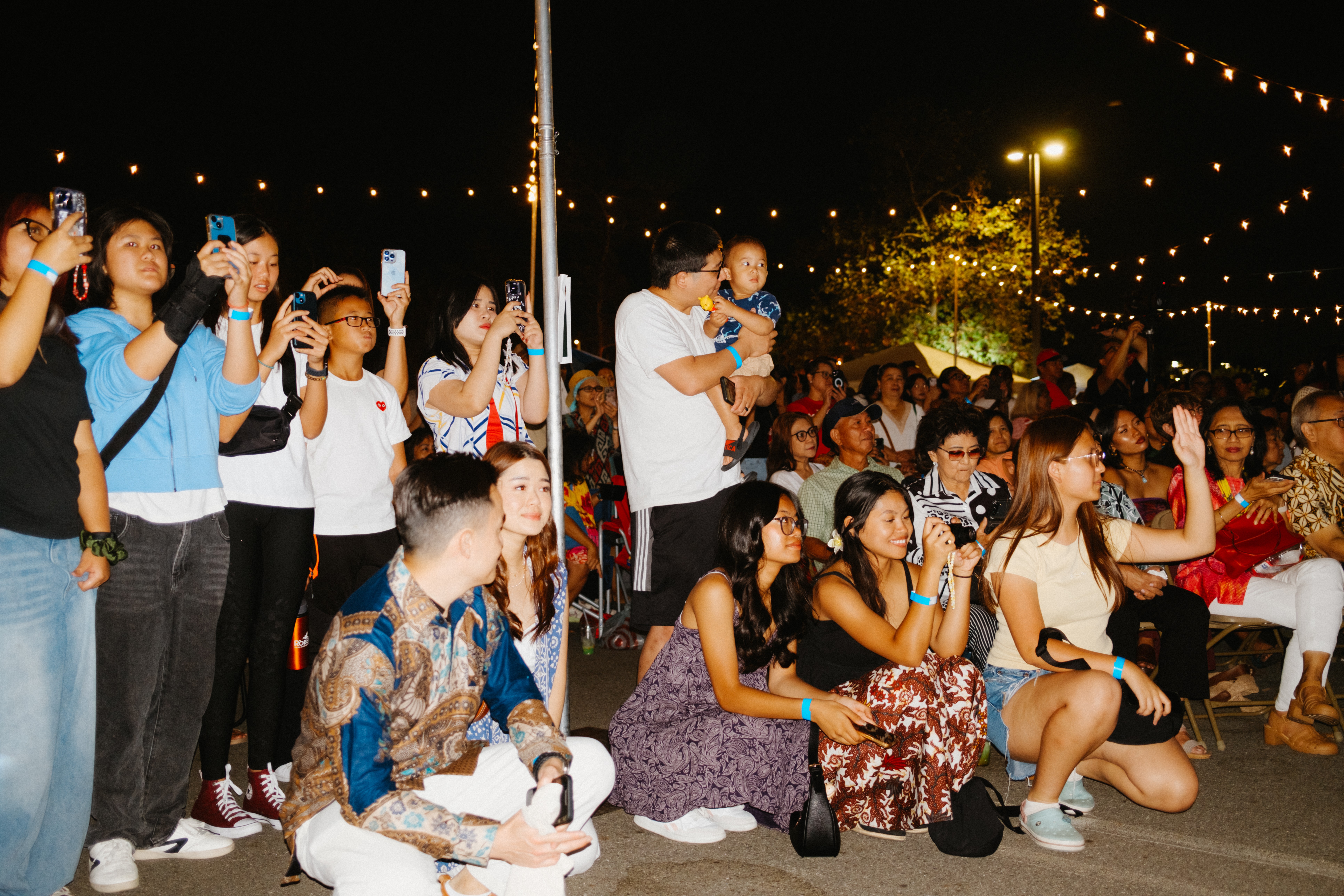 Group of people at an outdoor event at night, some standing and others sitting, with string lights overhead.