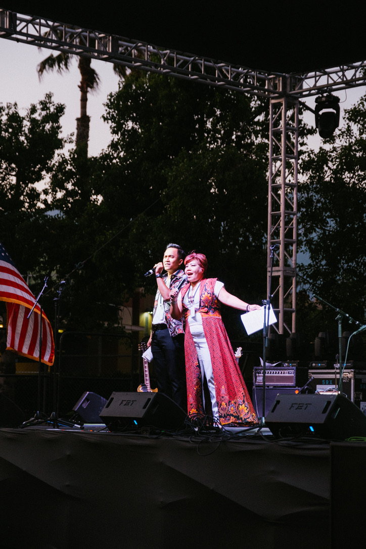 Two performers on stage, one singing and the other in a costume, with trees and stage equipment in background.