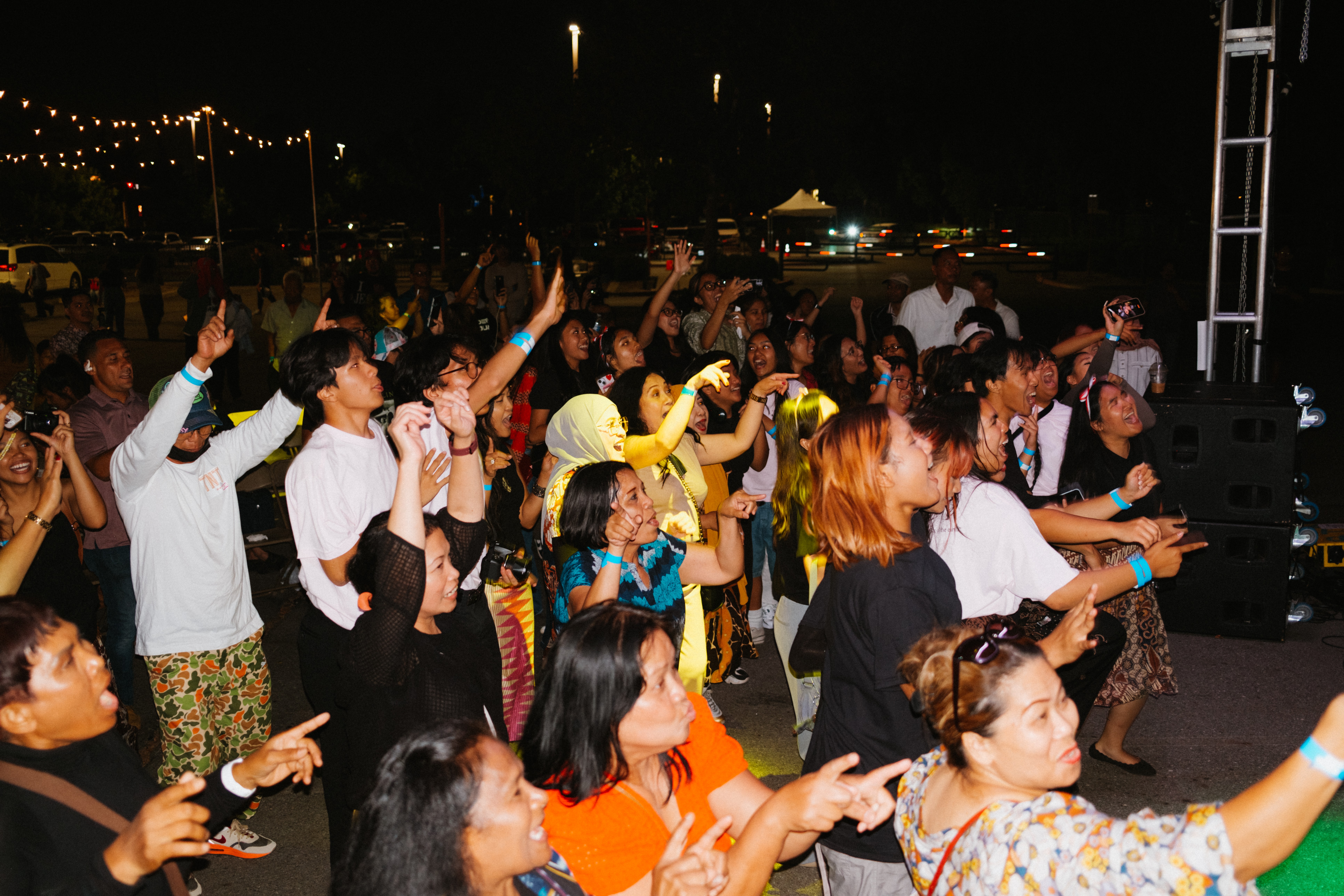 Crowd of people dancing and enjoying music outdoors at night with lights and stage equipment visible.