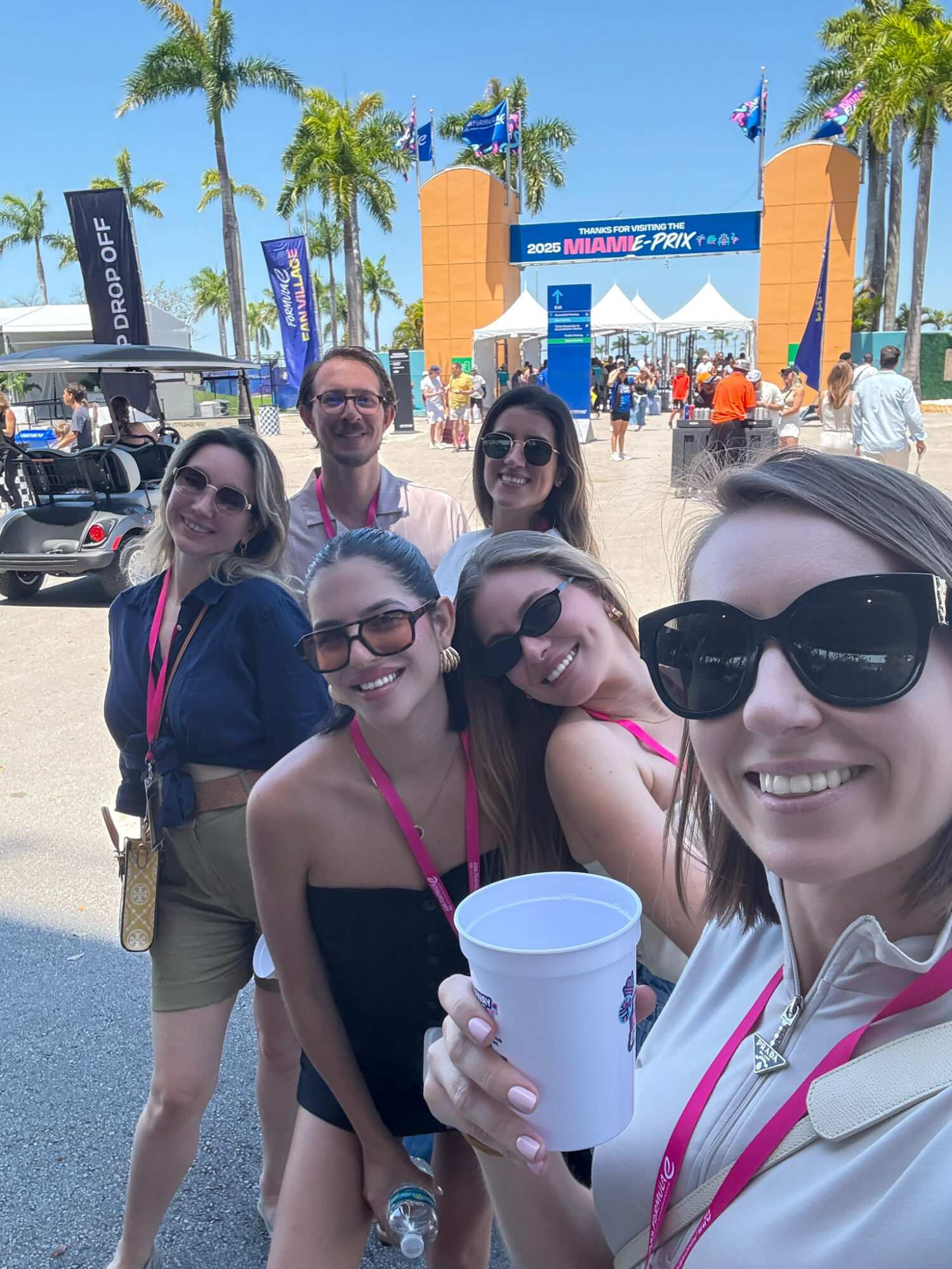 Group of six people smiling at an outdoor event with palm trees and a large archway in the background.