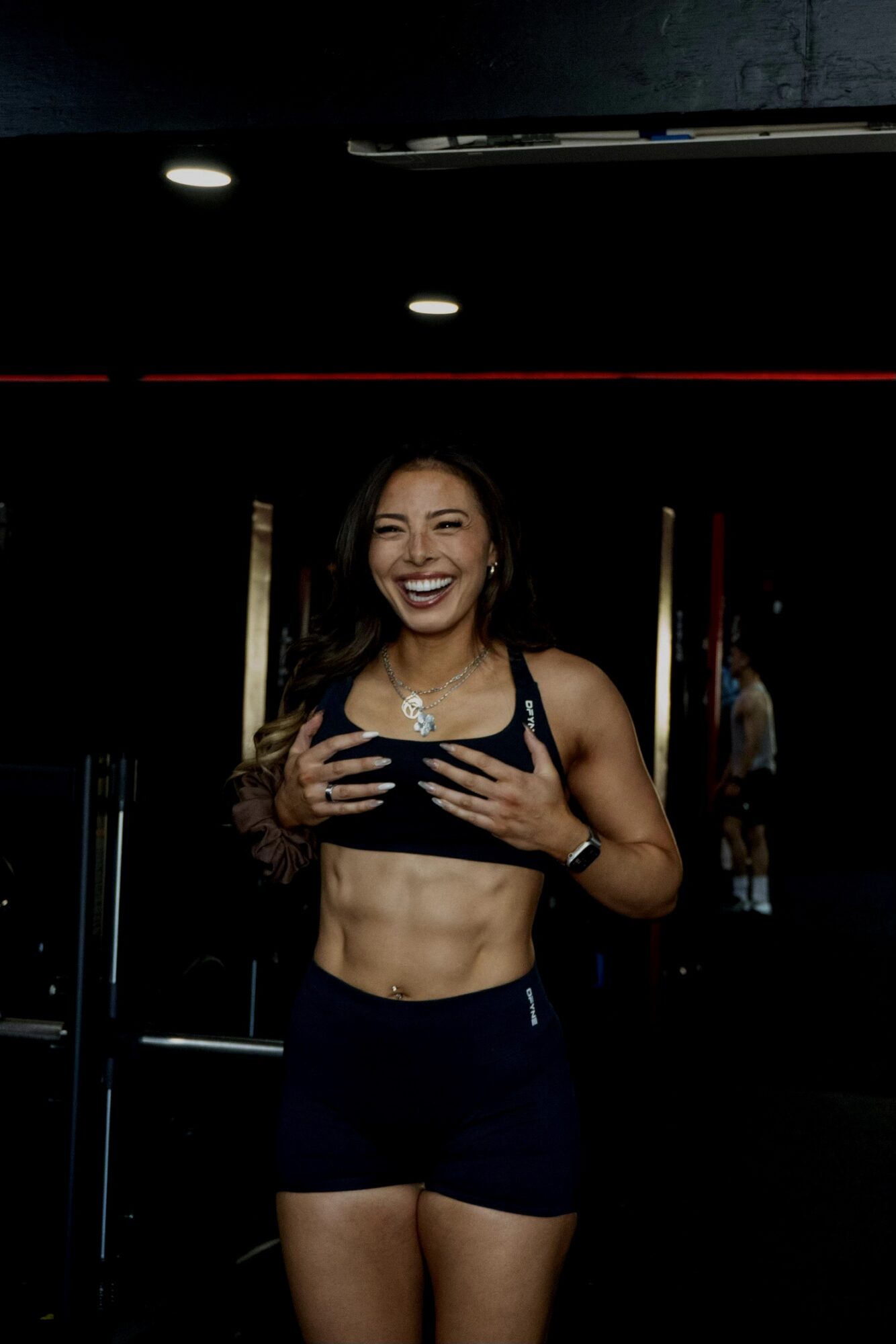 Smiling woman in sports bra and shorts taking a mirror selfie in a gym.