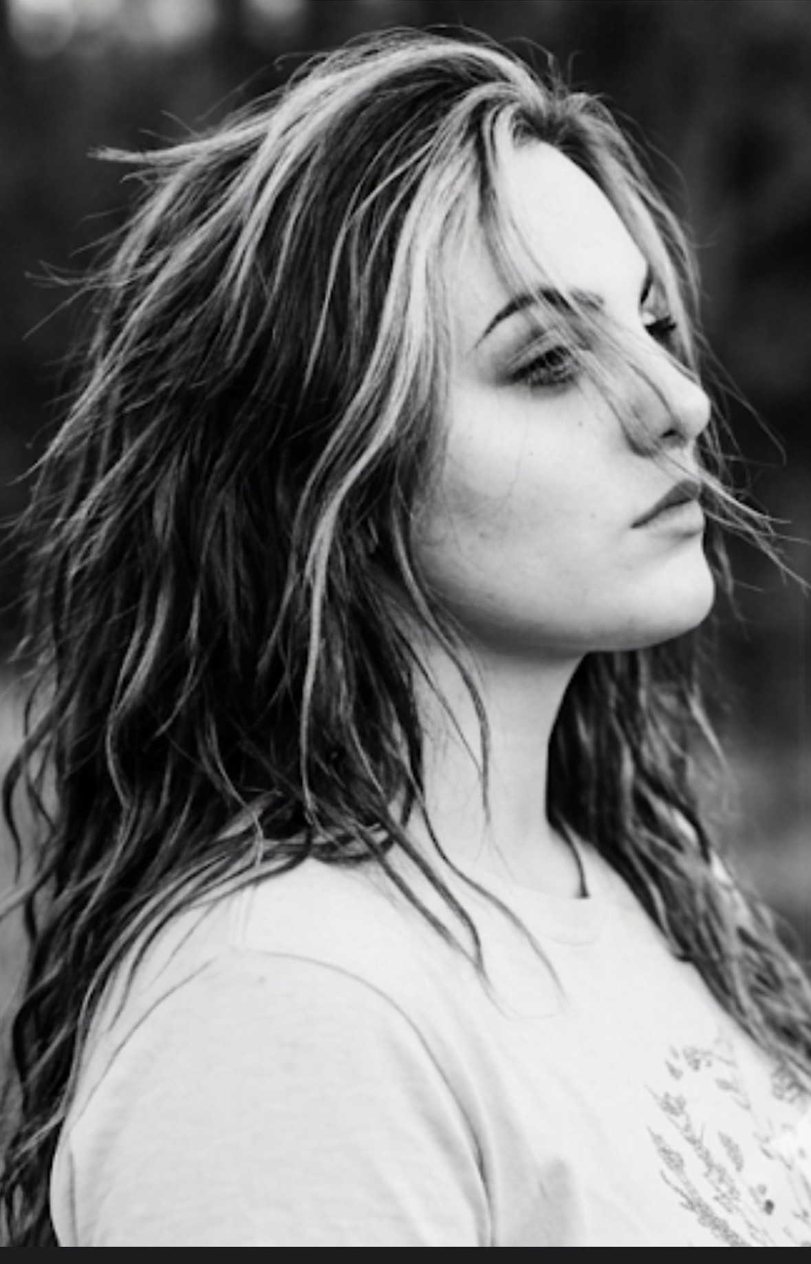 Side profile of a woman with long, wavy hair looking to the right, outdoors background, black and white photo.
