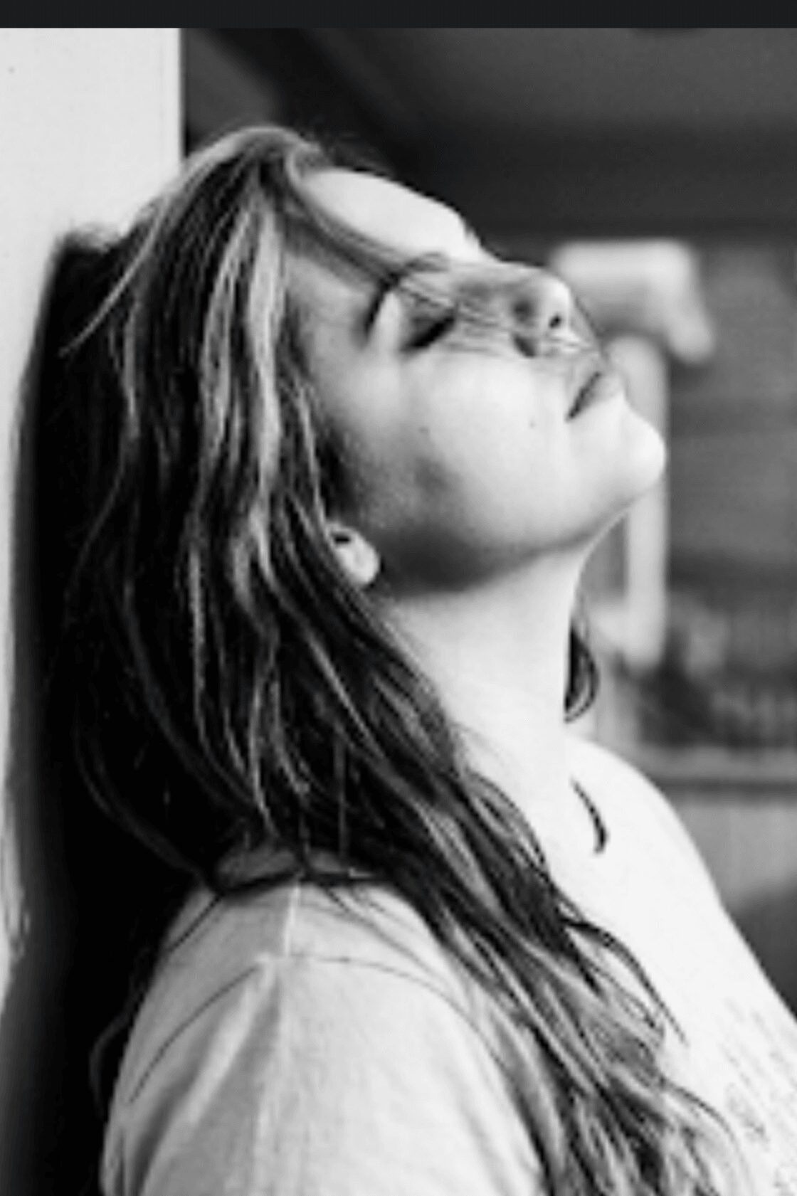Young woman with long hair leaning against a wall with eyes closed and head tilted back.