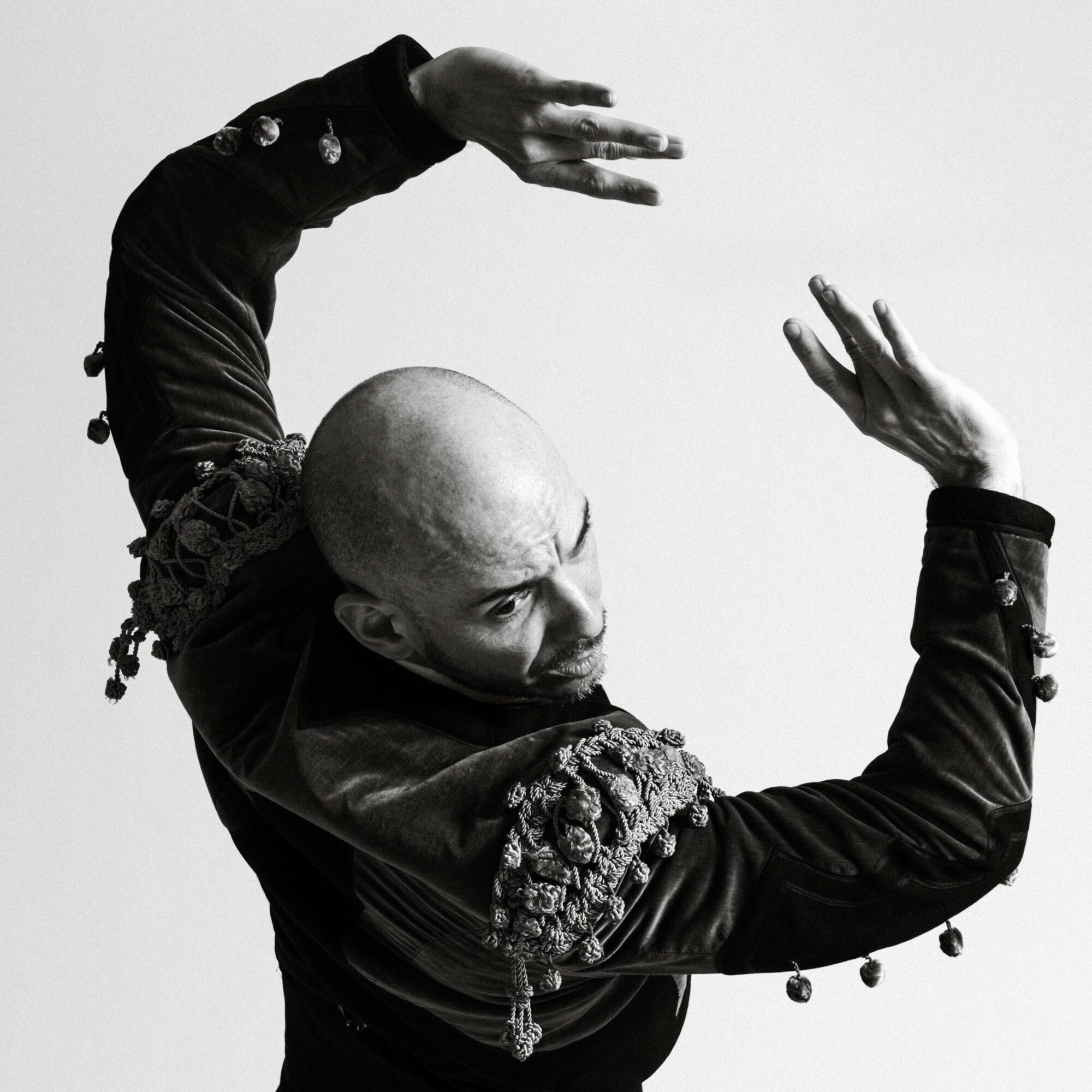 Person with bald head and elaborate sleeve pose, arms raised, in black clothing, against a plain background.