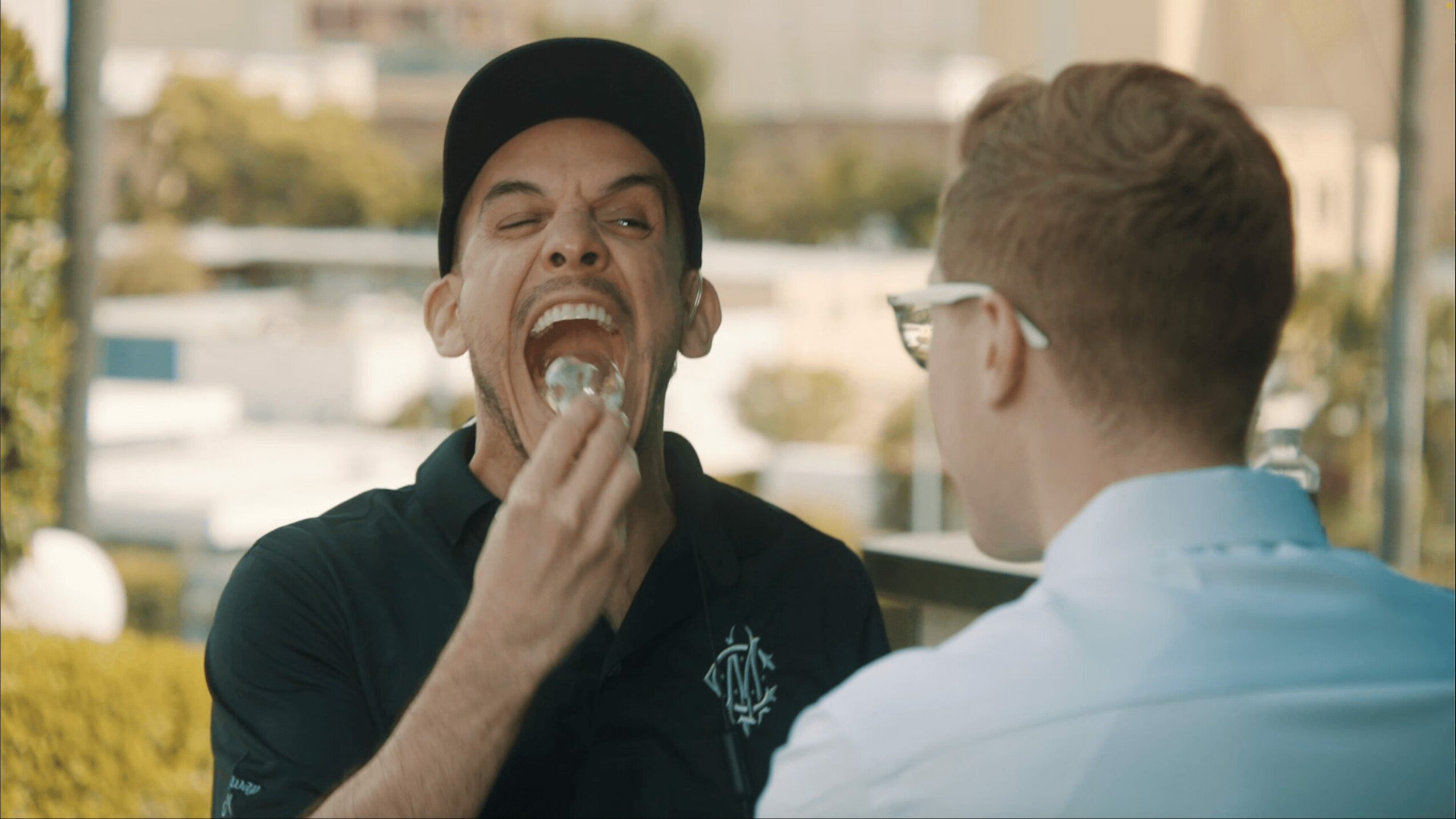 Two men engaged in conversation outdoors, one is eating, wearing a black cap, the other has glasses and light-colored shirt.