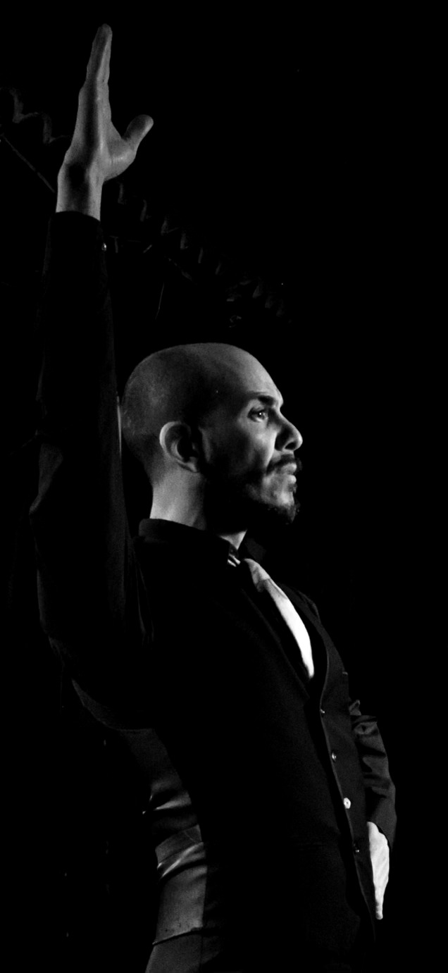 Man with beard and bald head raising his right hand, wearing a dark suit, against a black background.