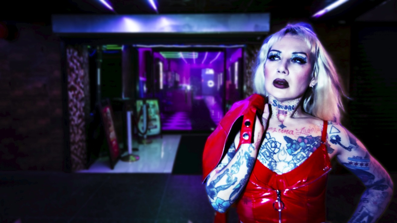 Person with blonde hair, tattoos, red outfit, and makeup in a dimly lit, colorful venue.
