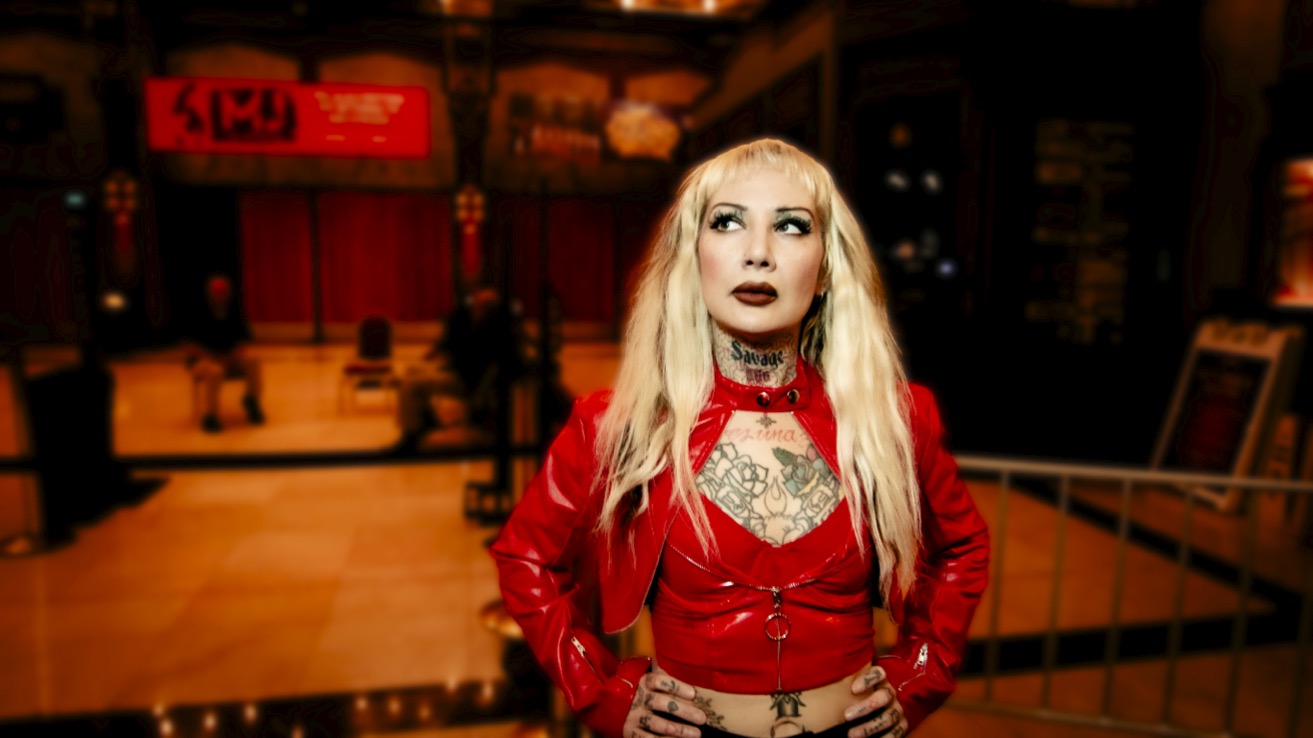 Woman with long blonde hair wearing a red jacket and choker, standing indoors with a bar or stage in background.