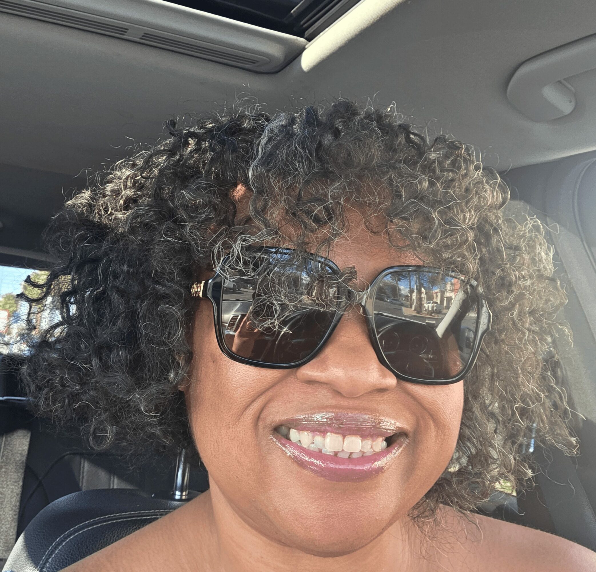 Woman with curly hair wearing sunglasses and smiling inside a car.