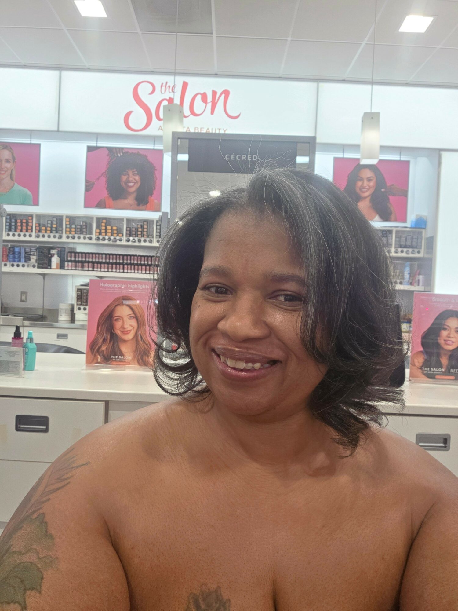 Woman with shoulder-length dark hair smiling in a salon with hair products and posters in background.