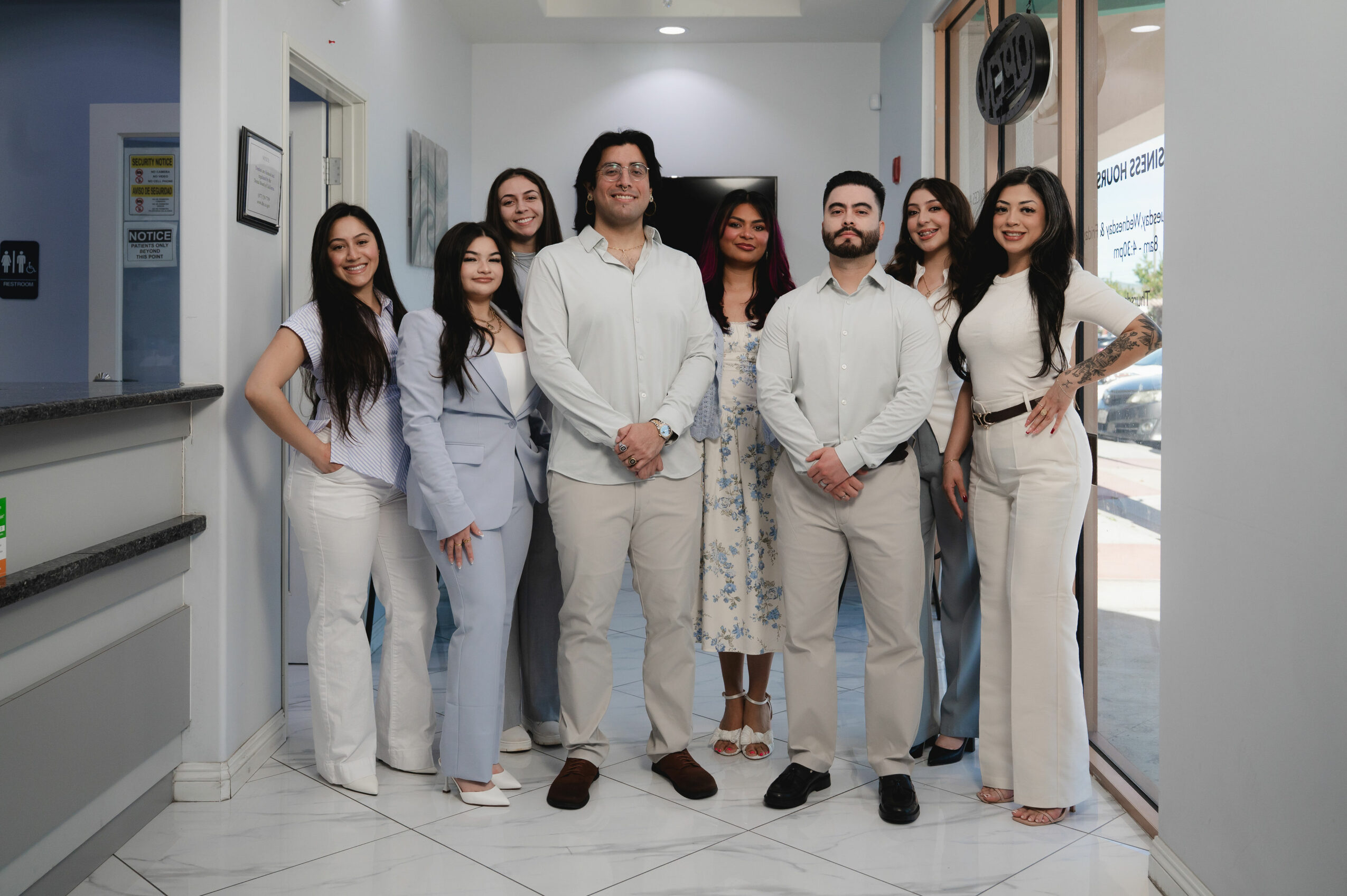 Group of eight people standing indoors, smiling, in a well-lit room with white walls and large windows.