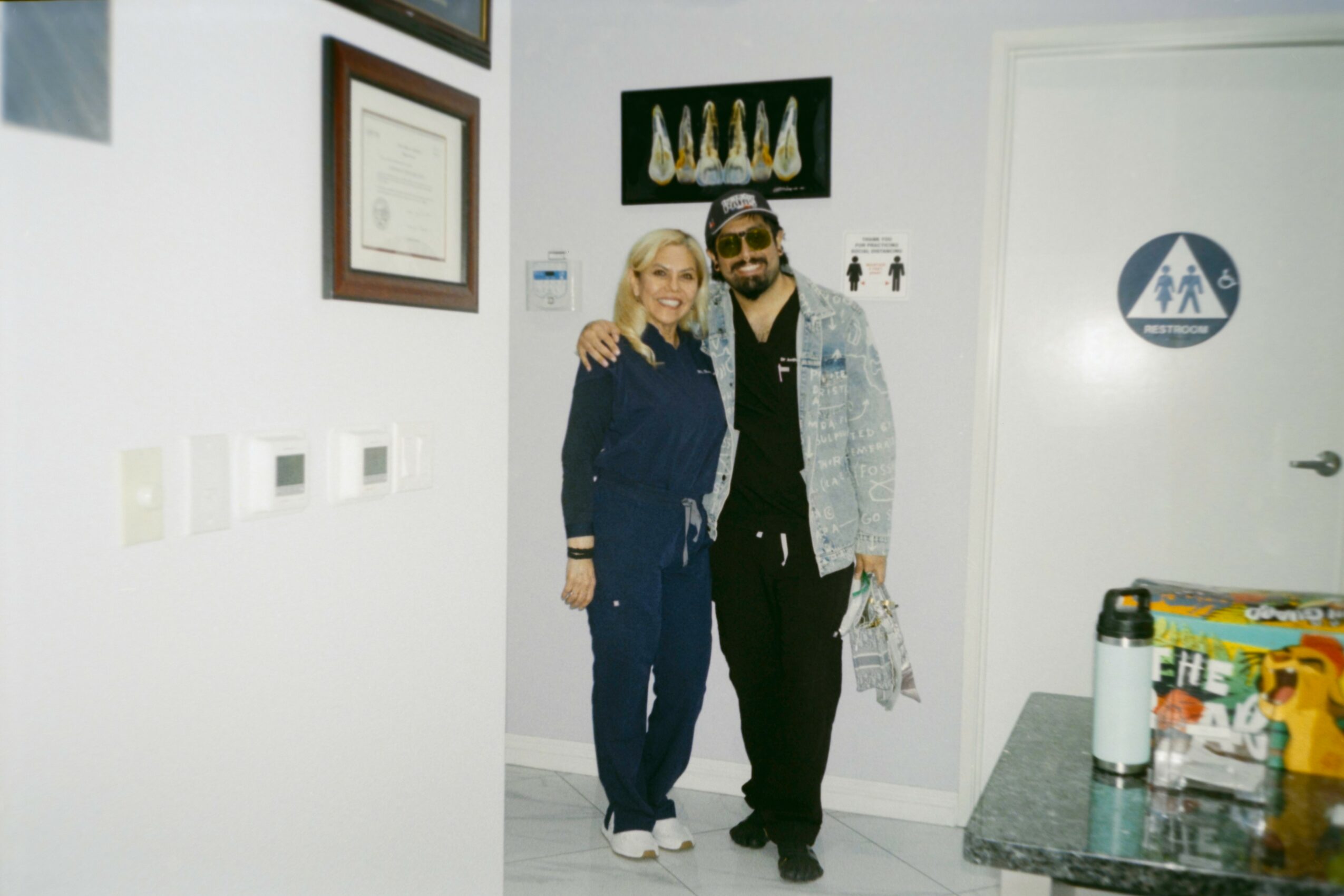Two people standing together in a room, one with arm around the other, smiling at the camera.