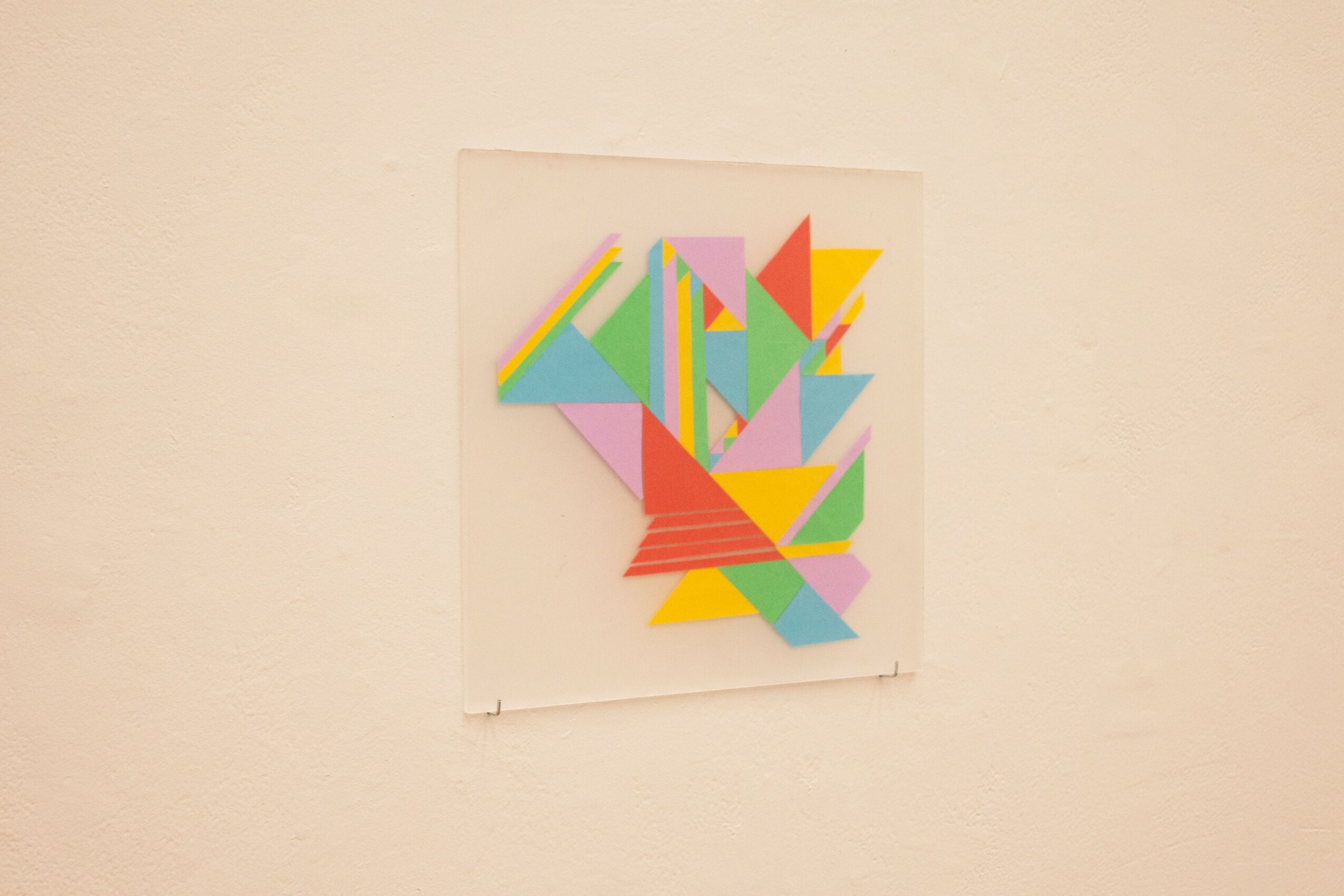 Colorful geometric abstract artwork on a white wall, featuring triangles and rectangles in pink, yellow, green, blue, and orange.