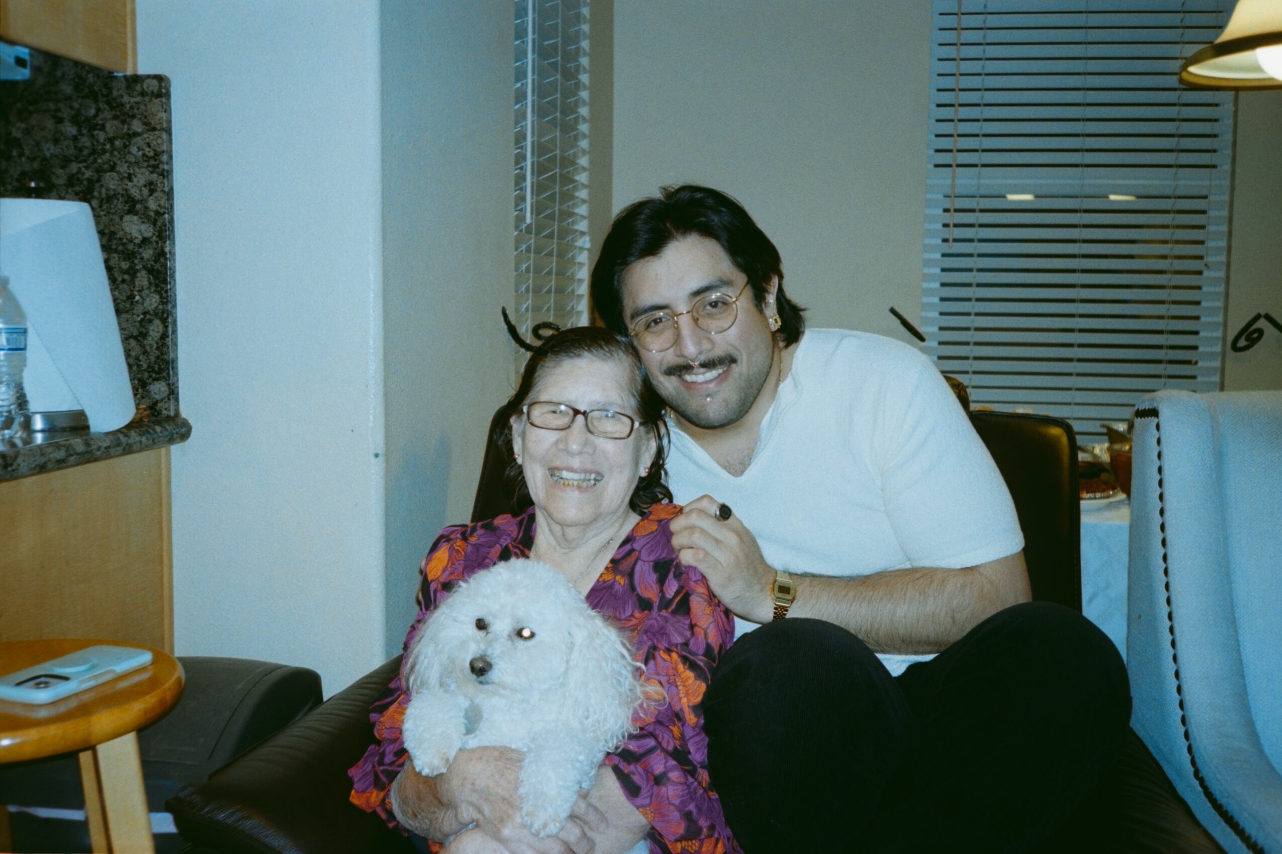 Two people smiling, sitting on a couch with a white fluffy dog, in a room with a window and blinds.