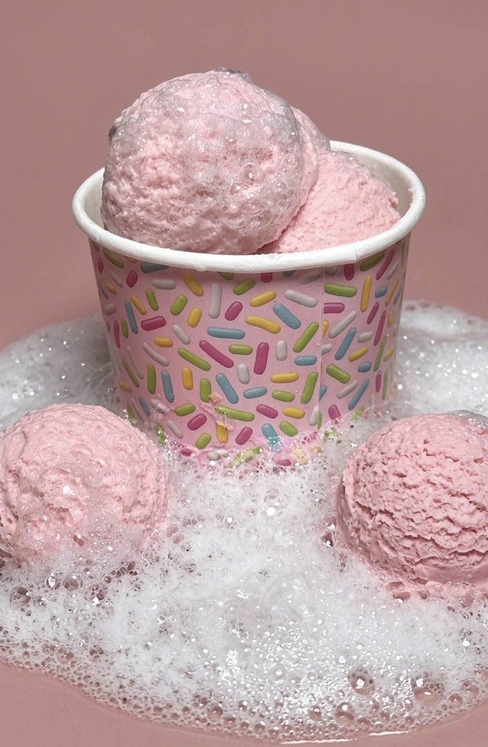 Pink bath bombs in a paper cup with colorful sprinkles design, surrounded by foam on a pink background.