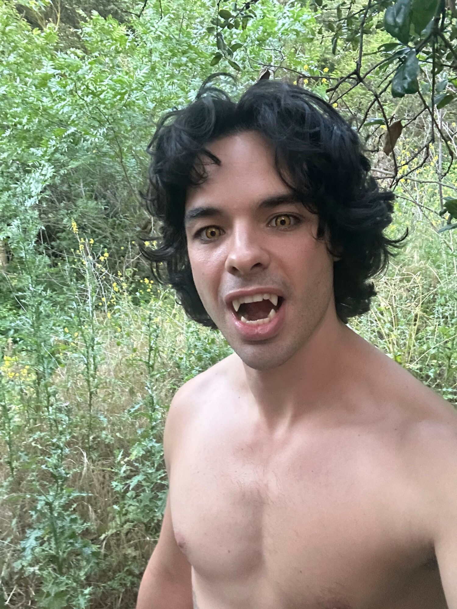 Young man with black curly hair and open mouth, shirtless, outdoors with green foliage background.