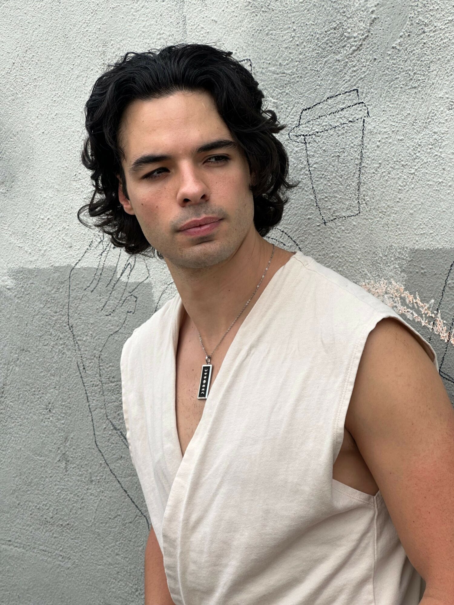 Young man with dark wavy hair and light sleeveless shirt standing against a textured wall.