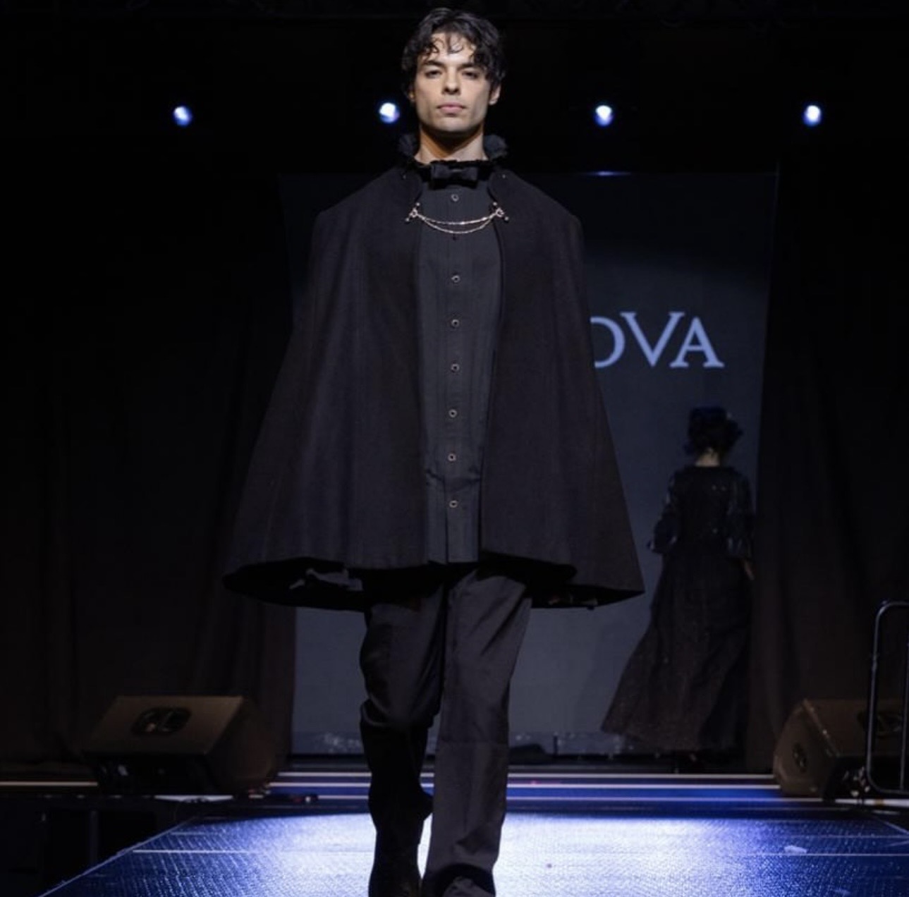 Male model walking on runway in dark clothing, stage with lights and a screen in the background.