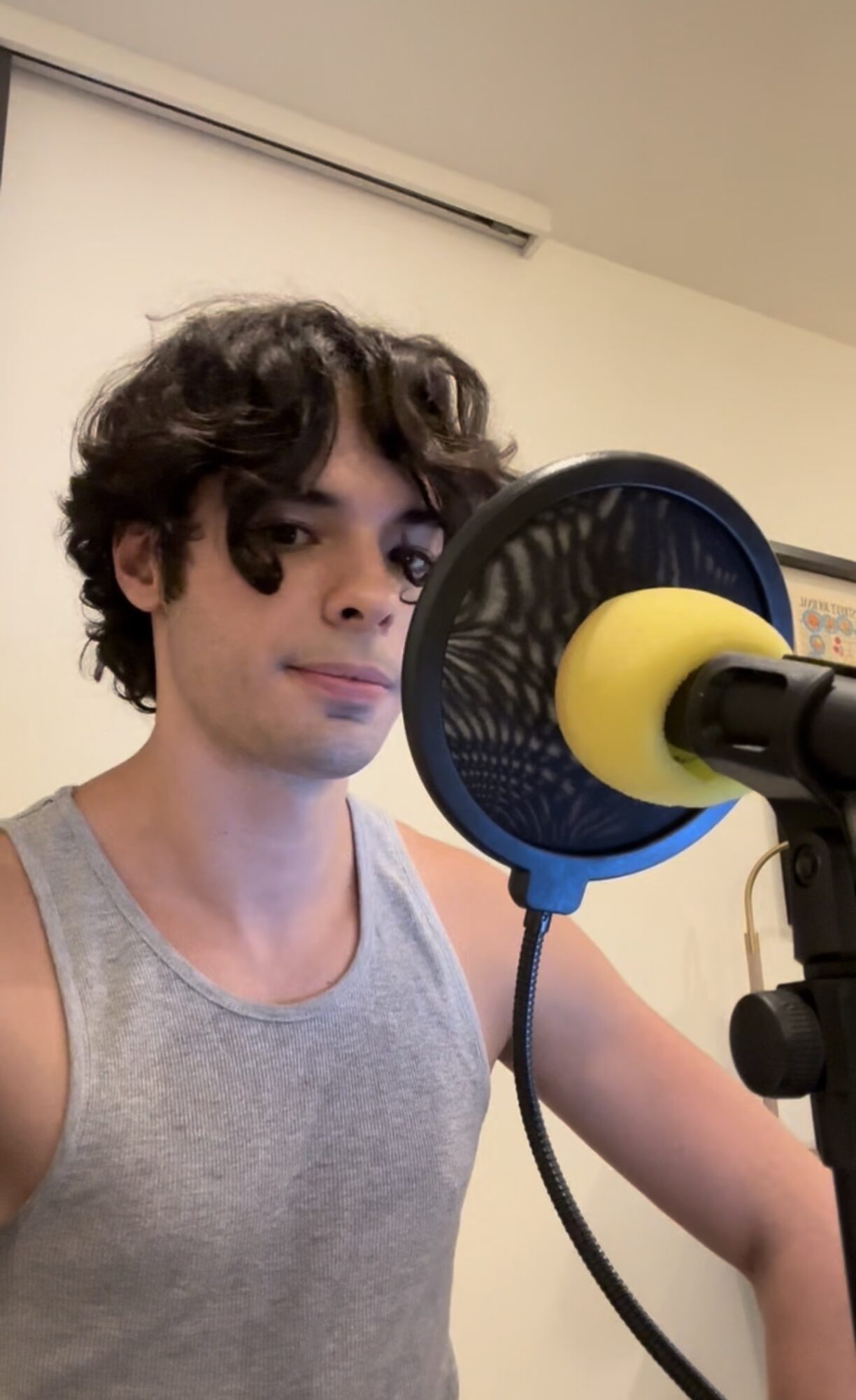 Young man with curly hair in a sleeveless shirt speaking into a microphone with a yellow pop filter.