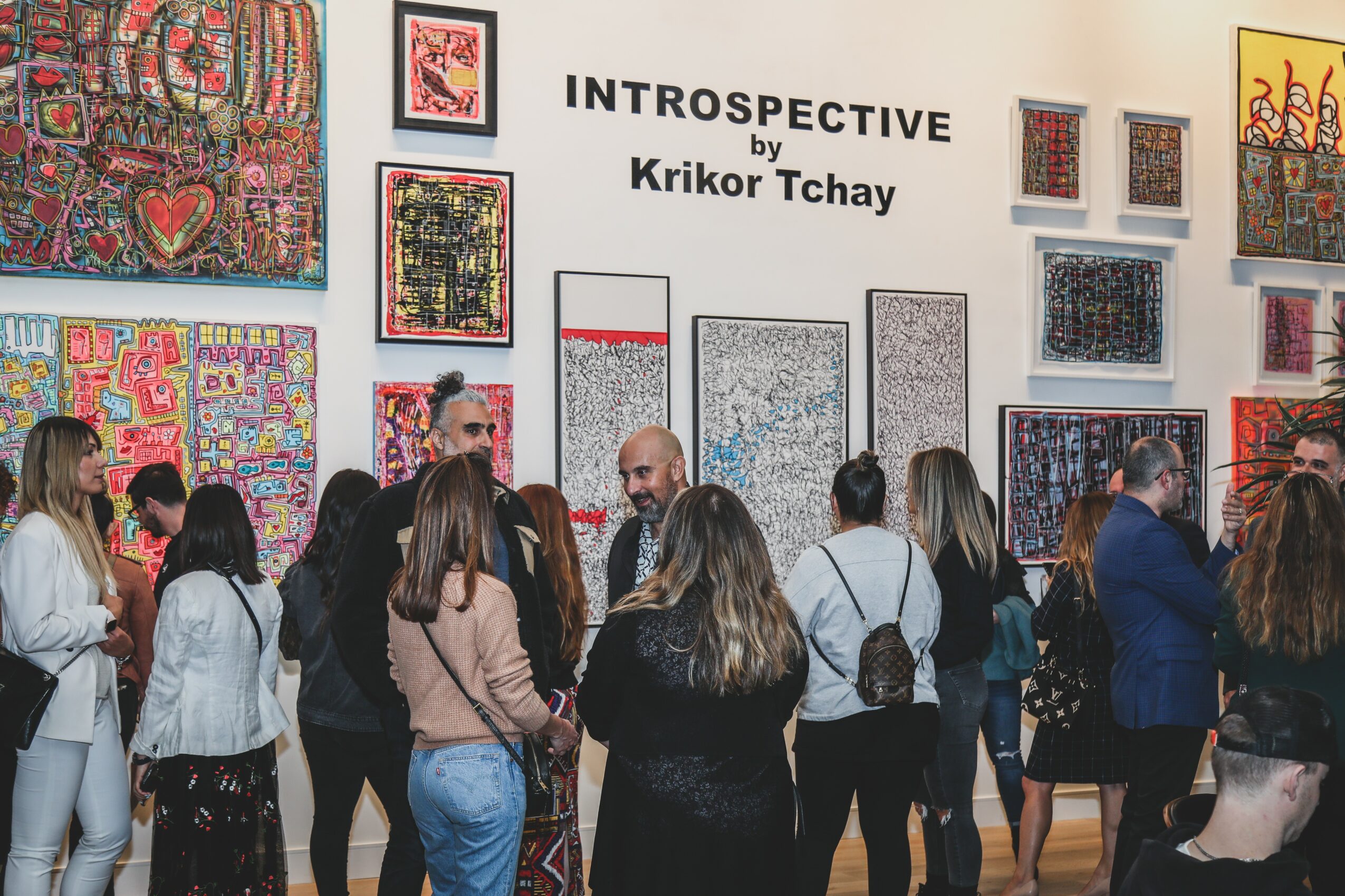 Group of people viewing colorful artwork at an art exhibition with a sign reading 'INTROSPECTIVE Krikor Tchay'.