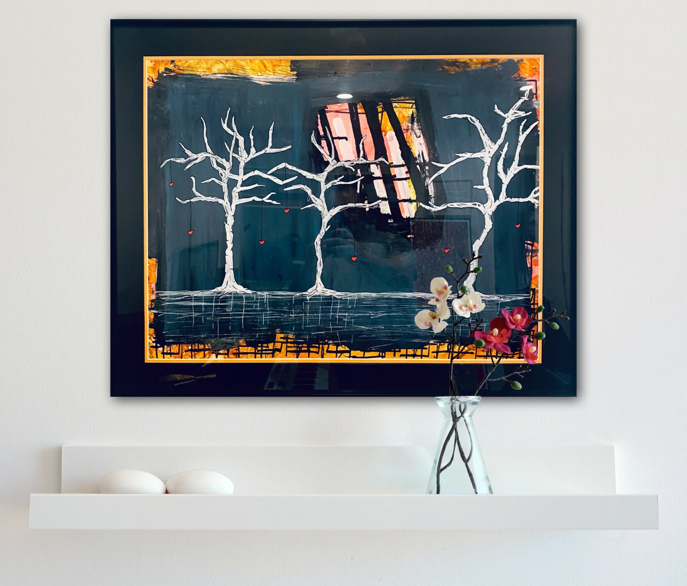 Framed artwork with white trees, books, and a small figure on a dark background, hanging above a white shelf with decorative objects.