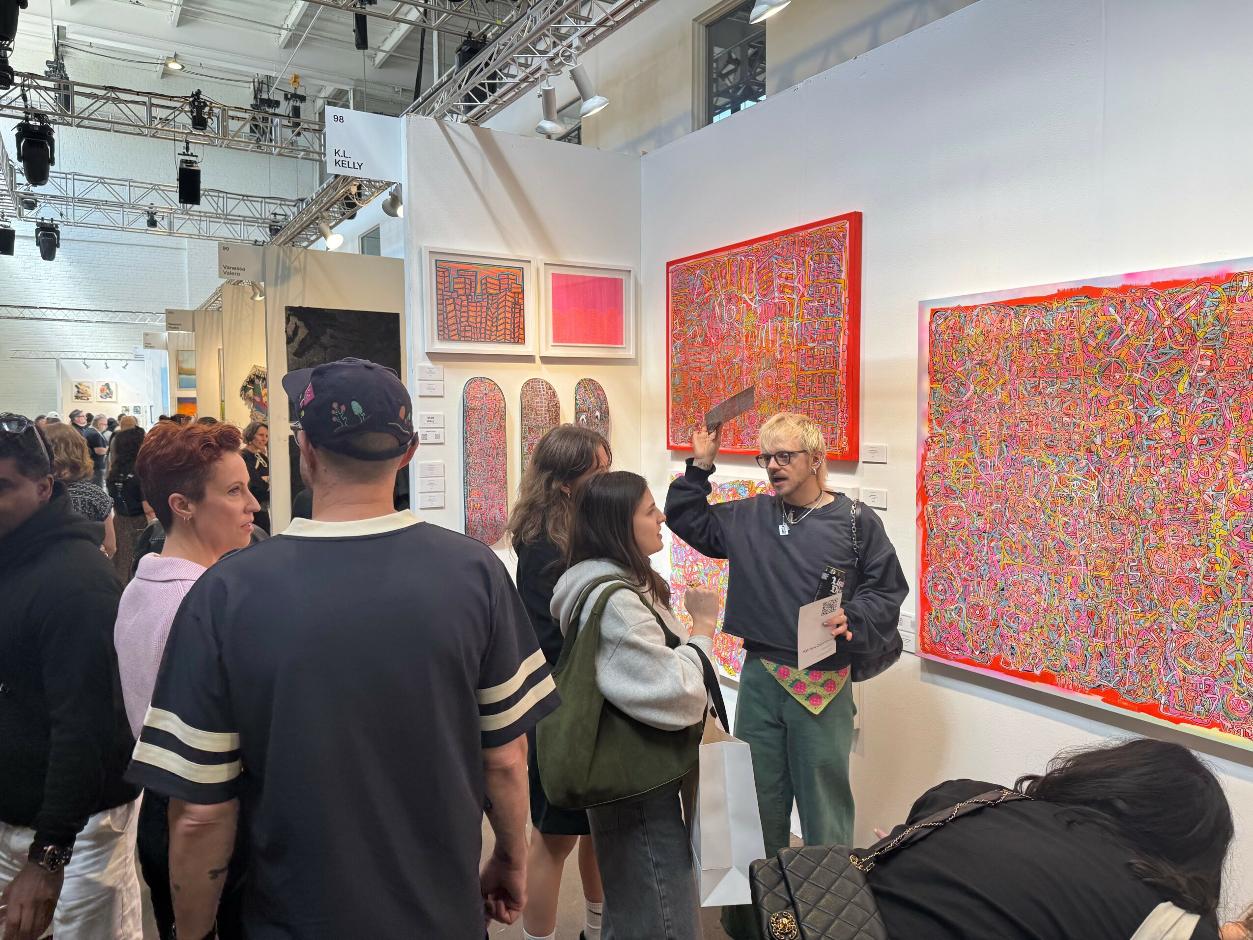 Group of people in an art gallery viewing colorful abstract paintings on white walls, with a guide explaining artwork.