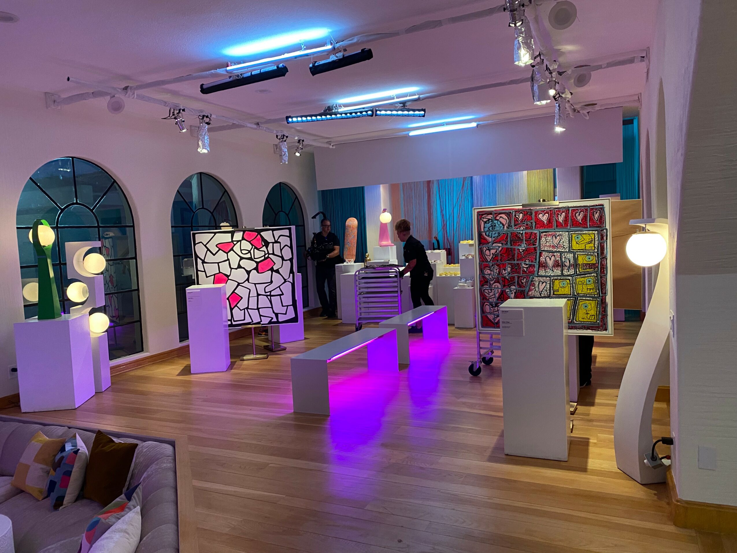 Art gallery with colorful paintings, white pedestals, and people viewing artwork, illuminated by ceiling lights and purple lighting.