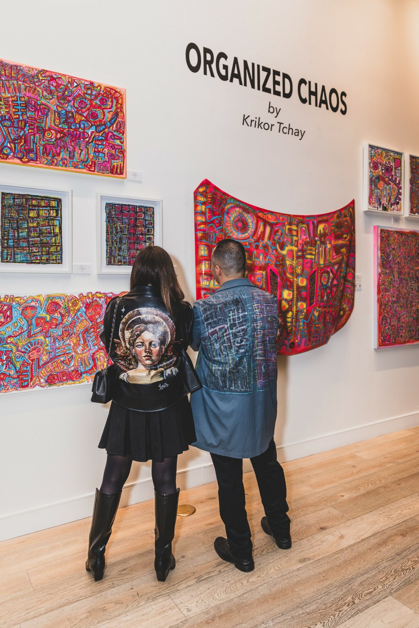 Two people observe colorful textile art pieces on a white gallery wall titled 'Organized Chaos.'