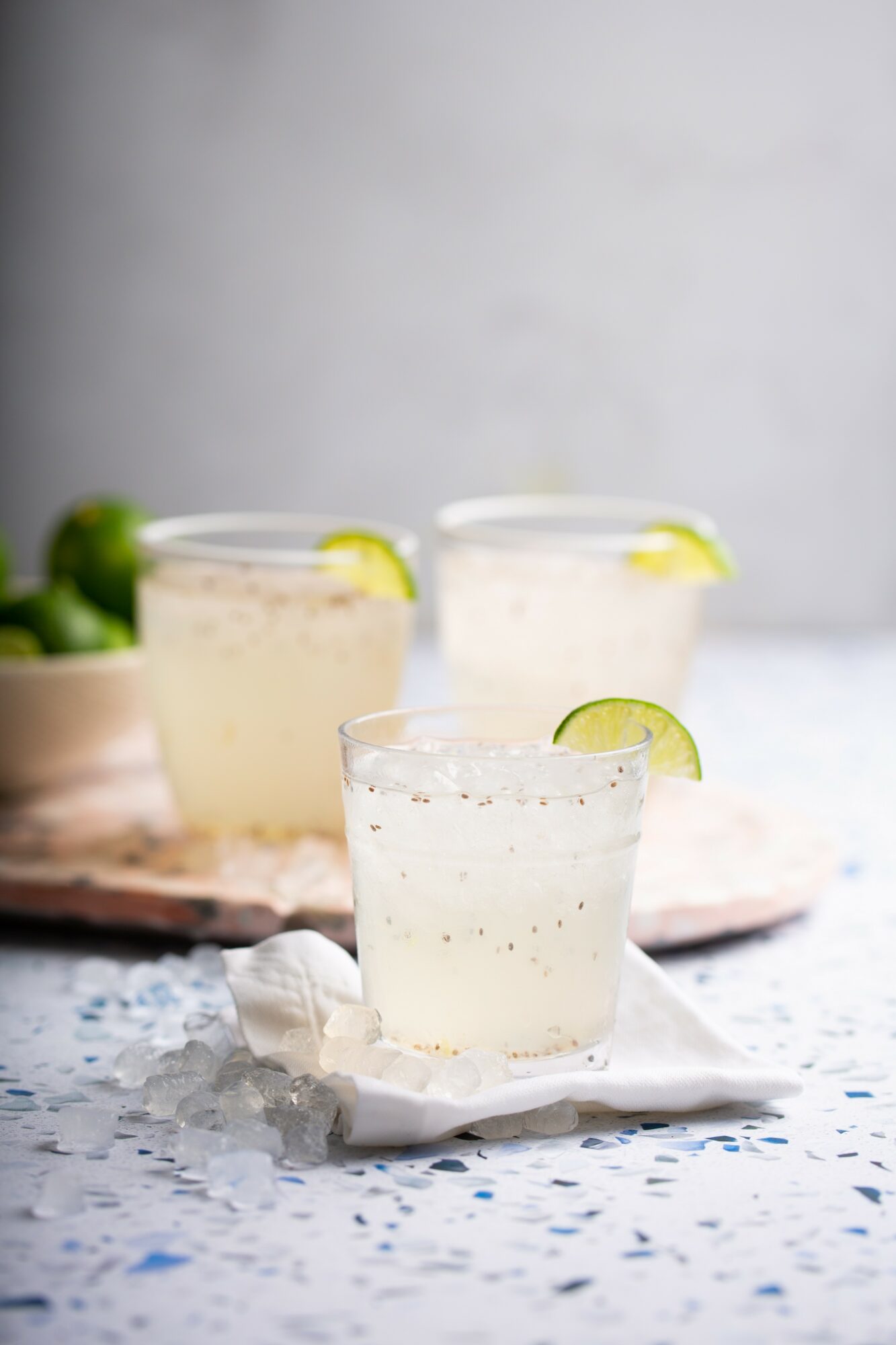Three glasses of lime-infused beverage on a white cloth, with a blurred background and scattered ice.
