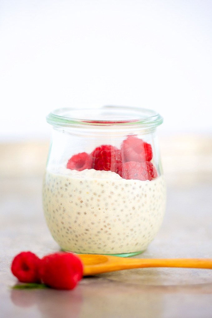 Glass jar filled with pudding topped with raspberries, with two raspberries and a yellow spoon in front.