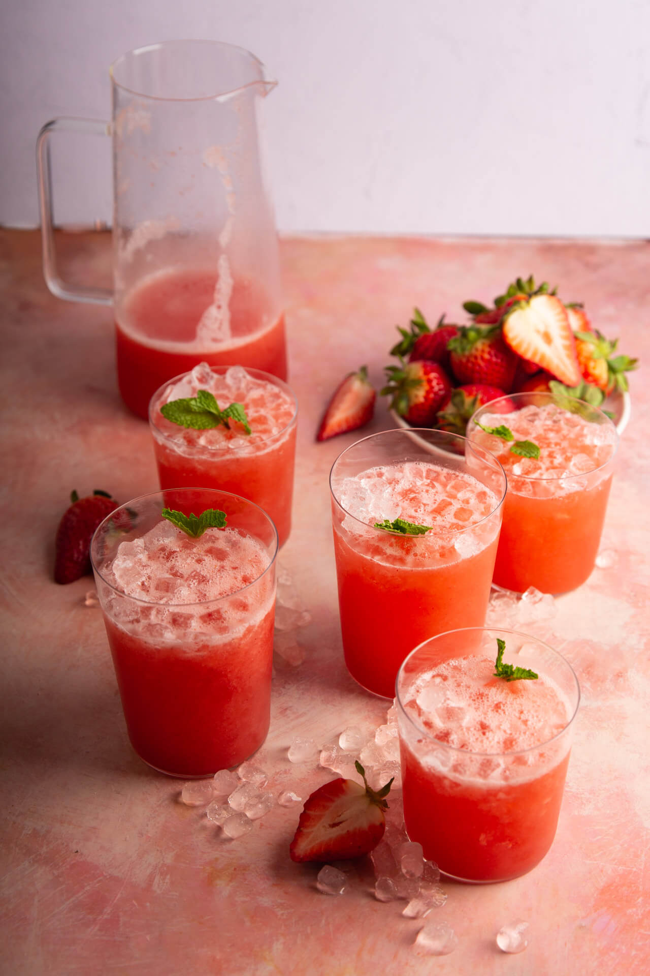 Six glasses of pink fruit juice with ice, strawberries, and mint leaves on a pink surface, with a pitcher in the background.