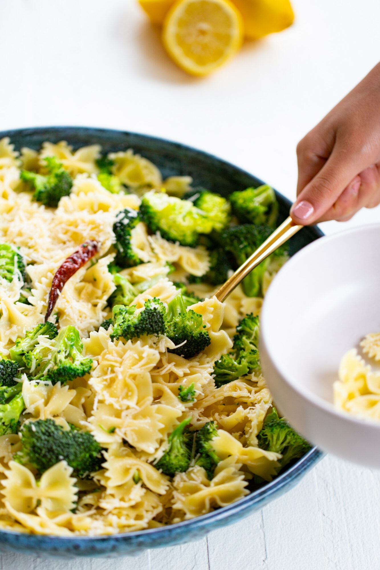 Hand holding a fork over a bowl of pasta with broccoli and grated cheese, lemon in background.