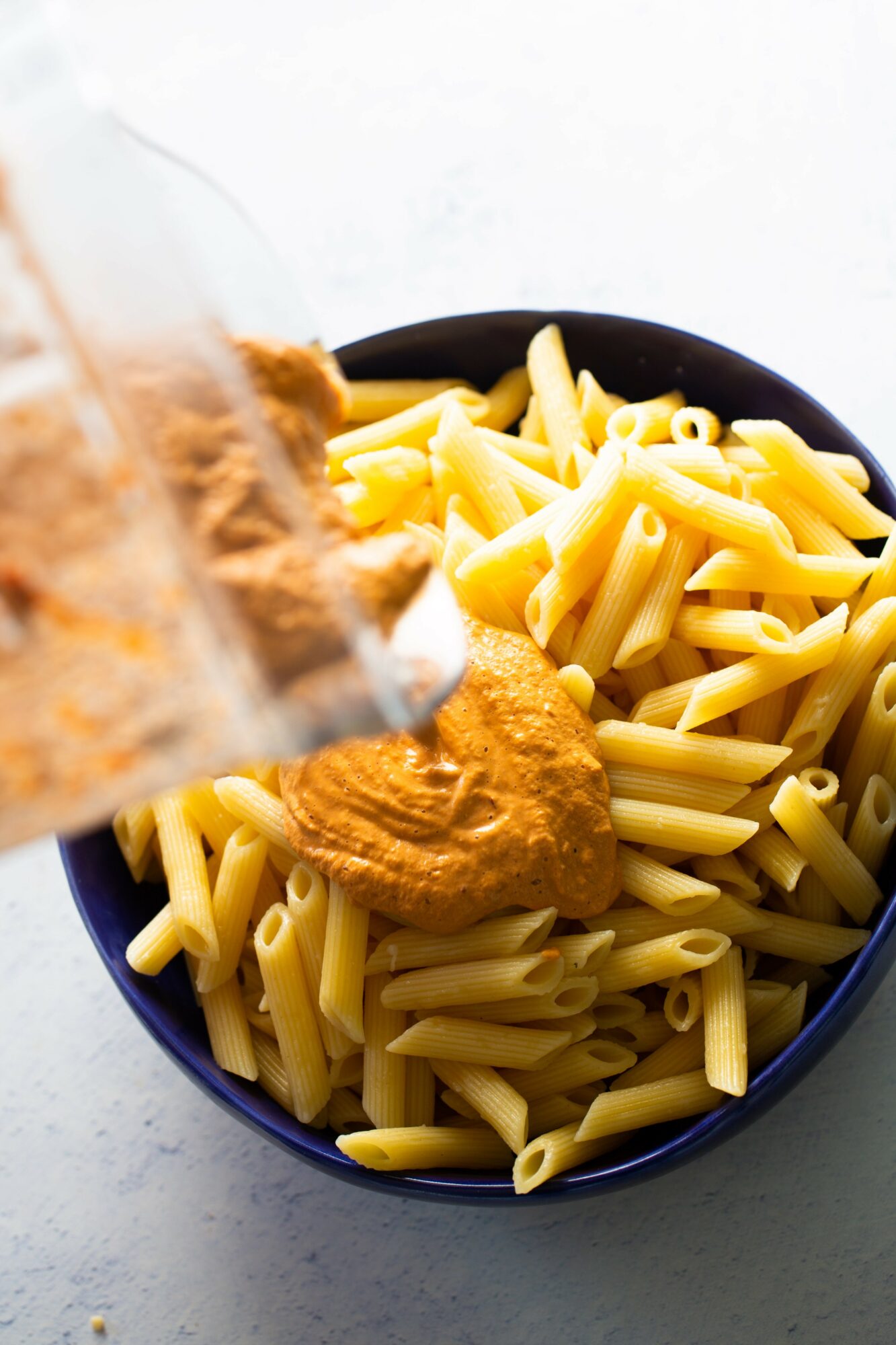 A bowl of cooked penne pasta with a dollop of sauce, with a spoon pouring sauce over it.