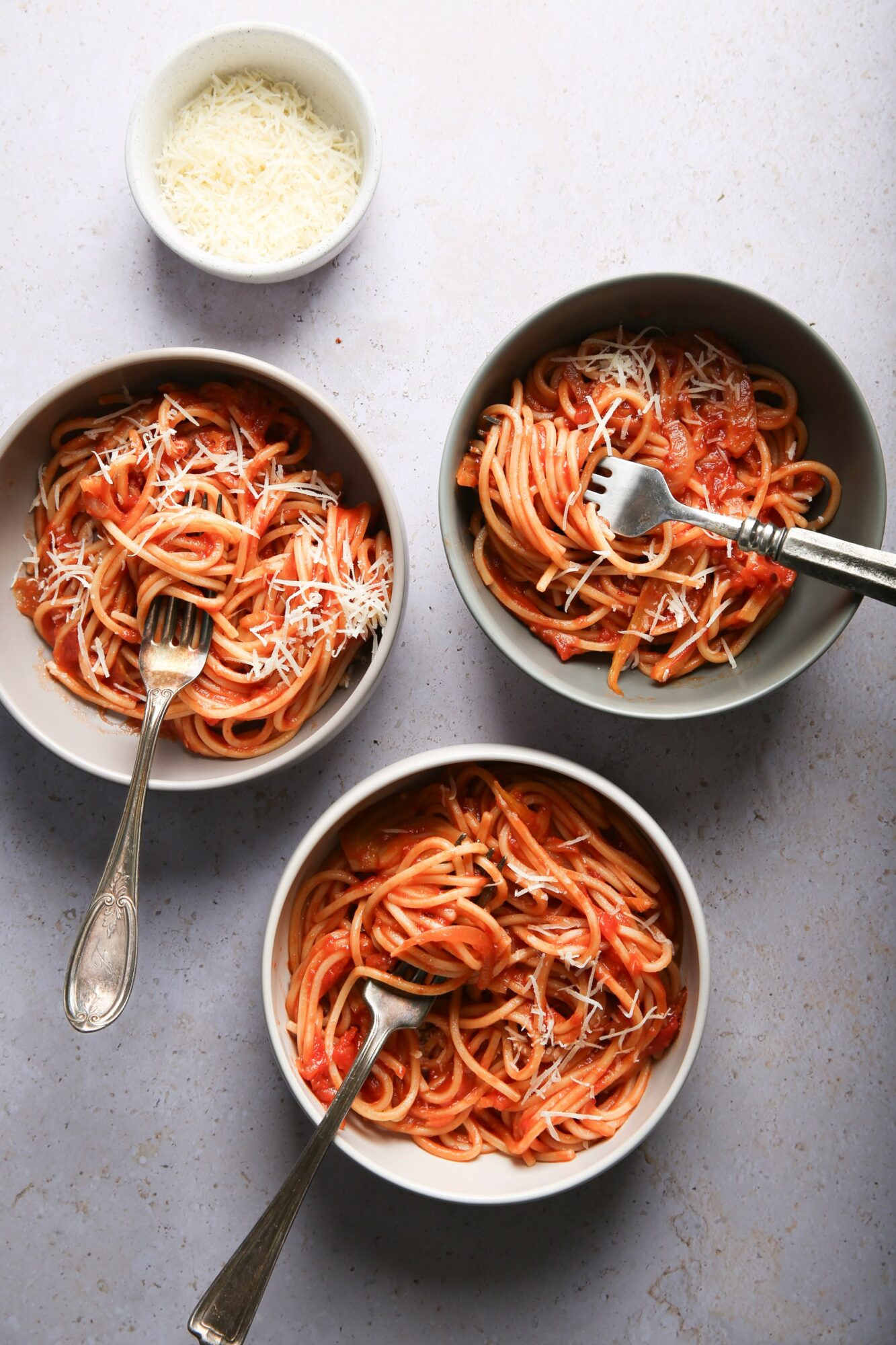 Three bowls of spaghetti with grated cheese on a light gray surface, one bowl with a fork, one with a fork and spoon.