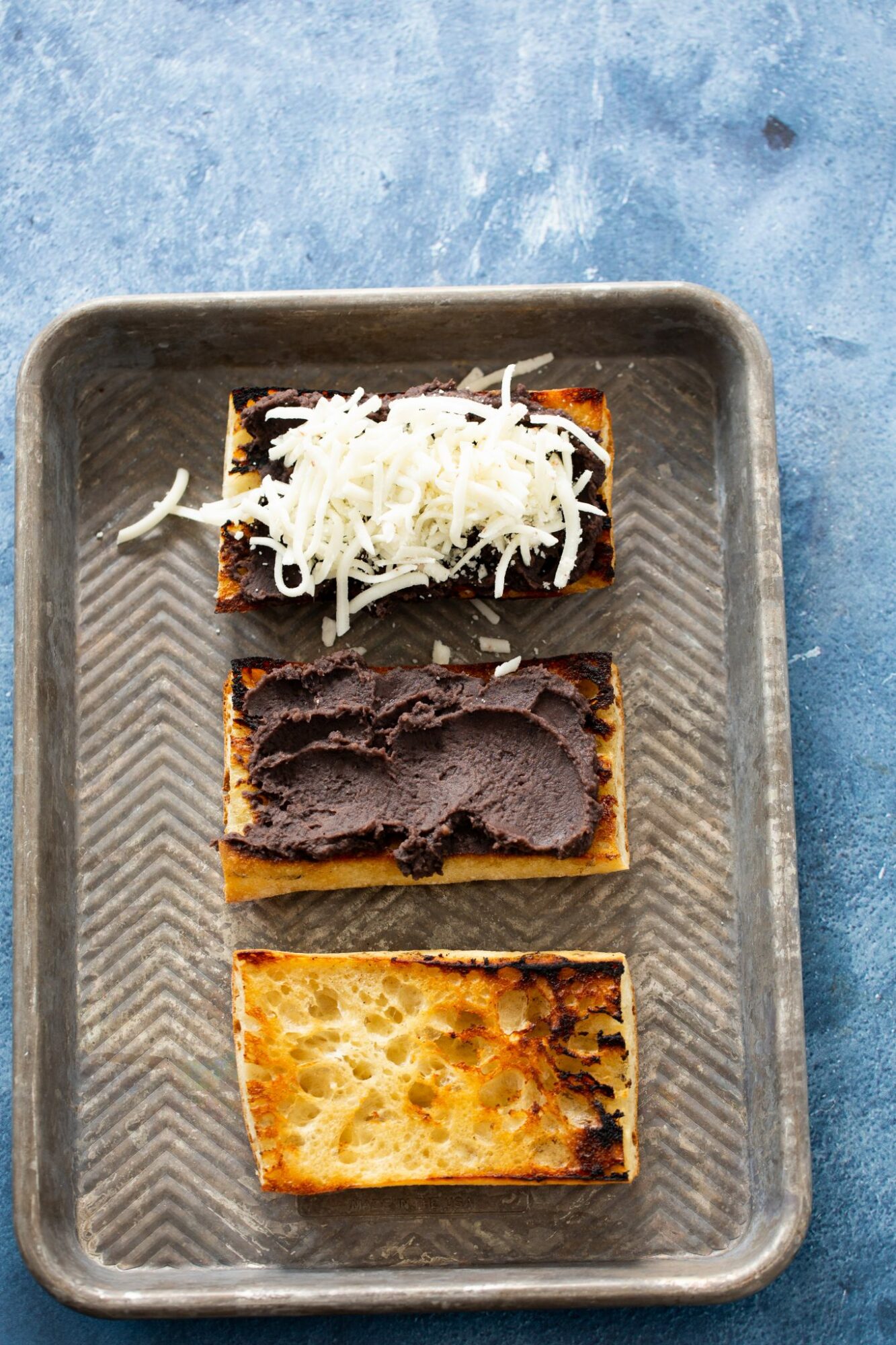 Three rectangular pieces of toast with toppings on a metal tray, on a blue surface.