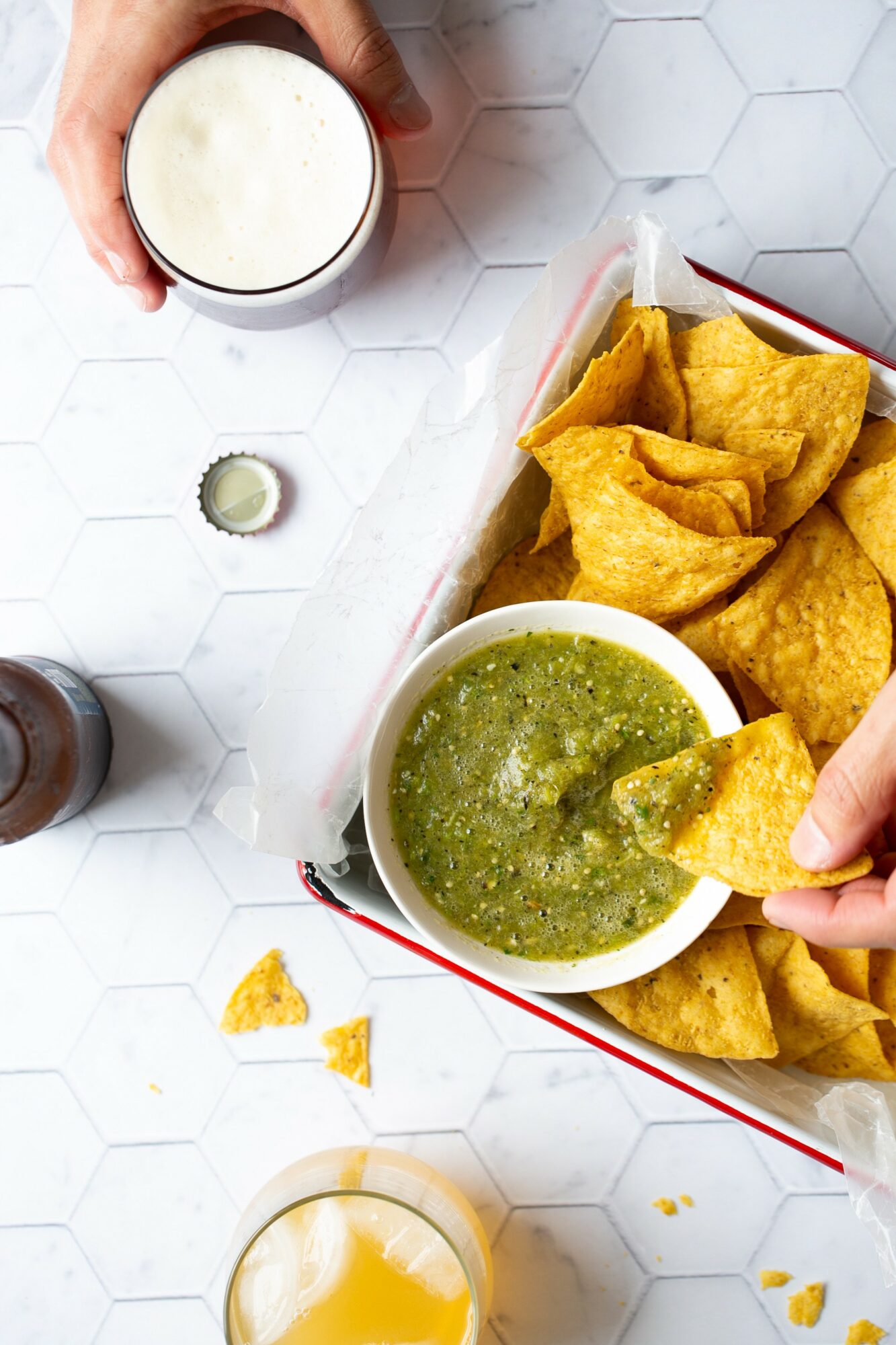 Hand dips tortilla chip into green salsa with chips in a basket on a white hexagonal tile surface.