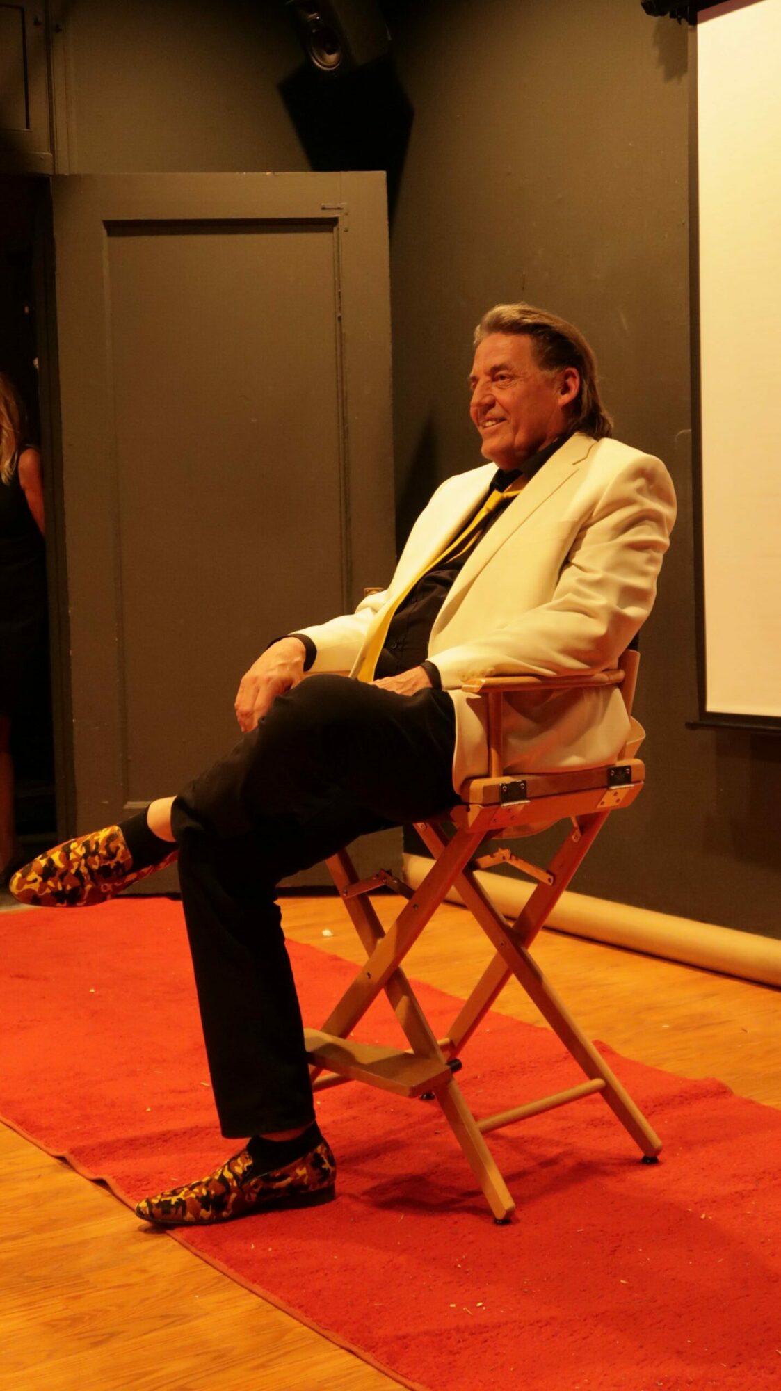 Person sitting on a wooden chair on stage, facing sideways, with a large screen behind them.