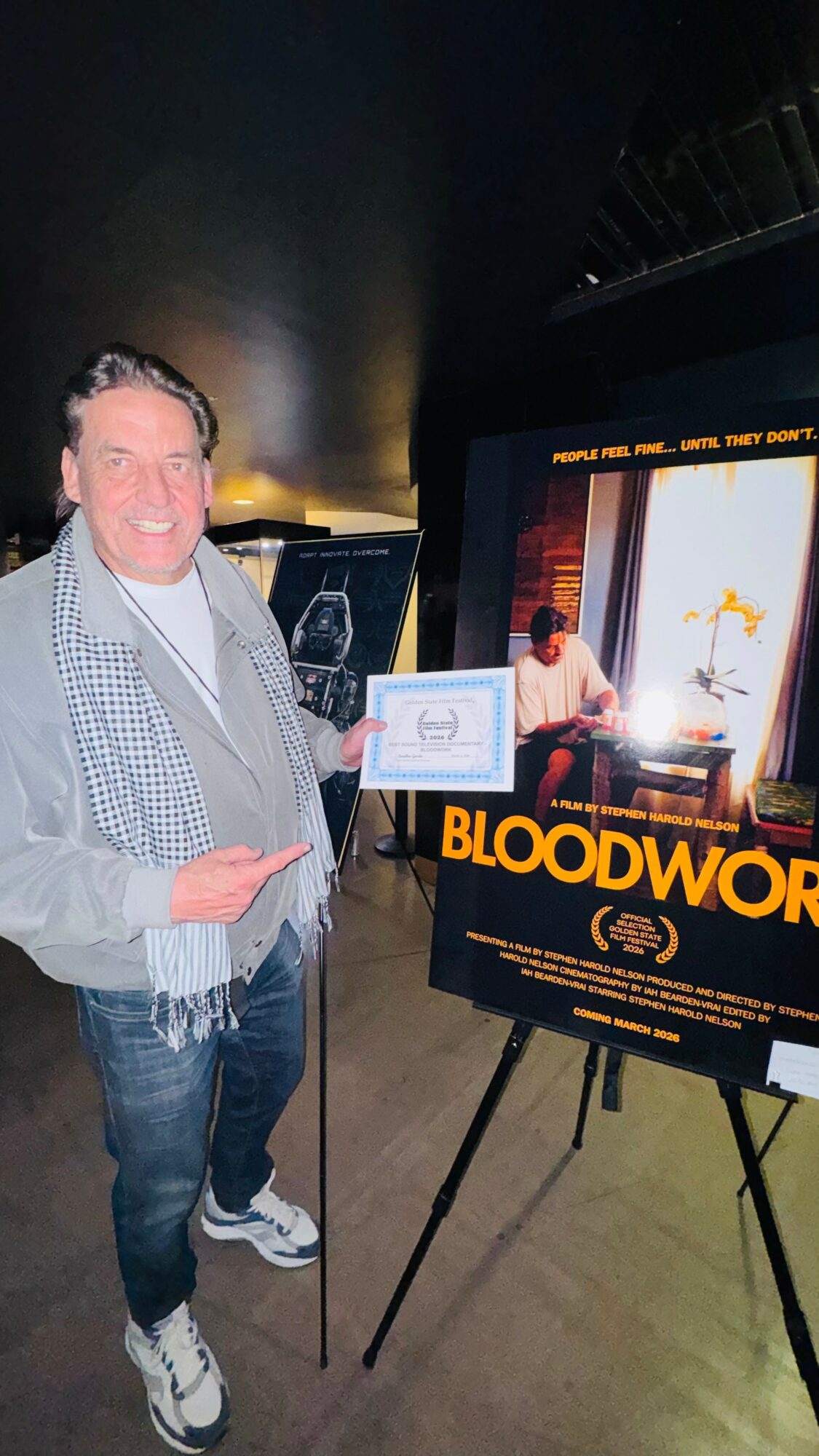 Man smiling holding a certificate next to a movie poster display for 'Bloodword'.